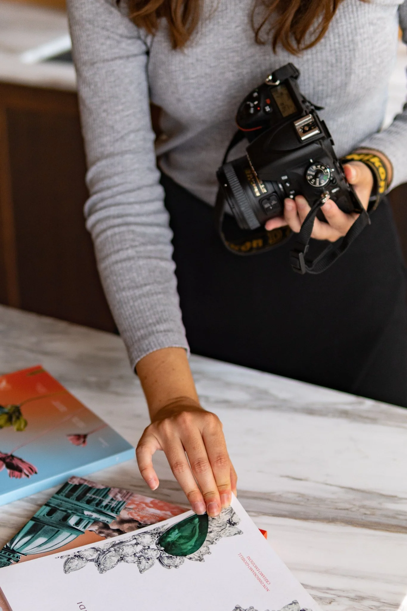 A woman holding a camera and flipping through pages of a magazine or catalog on a table.