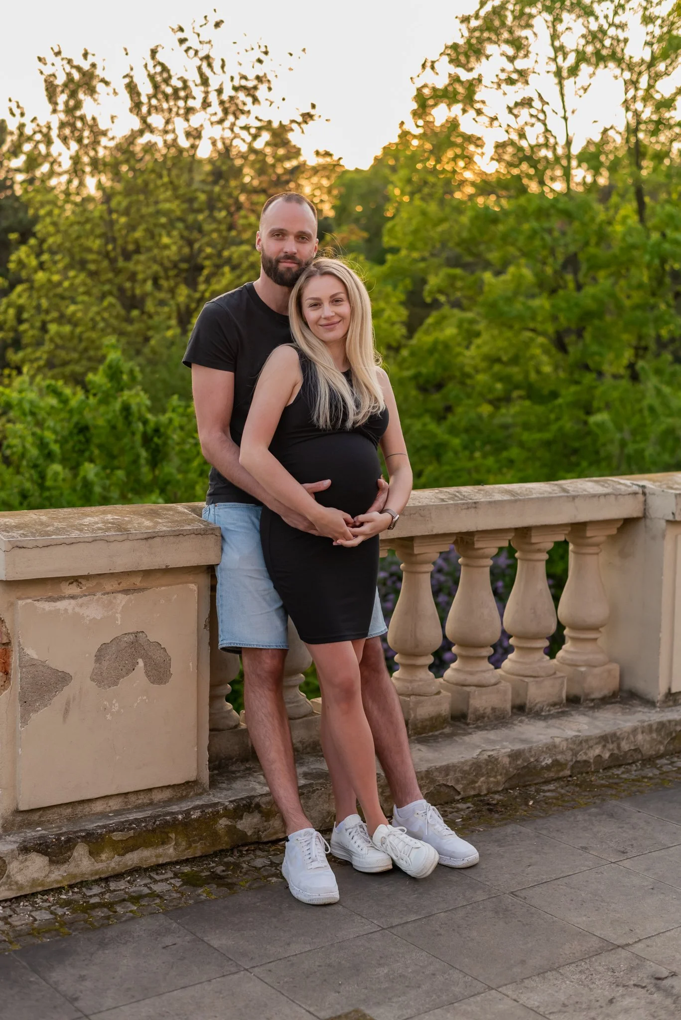 A couple standing outdoors on a terrace with a stone railing, with green trees in the background at sunset. The woman is pregnant, wearing a black dress and white sneakers, while the man is wearing a black t-shirt, light blue shorts, and white sneakers, standing behind her with arms around her waist.