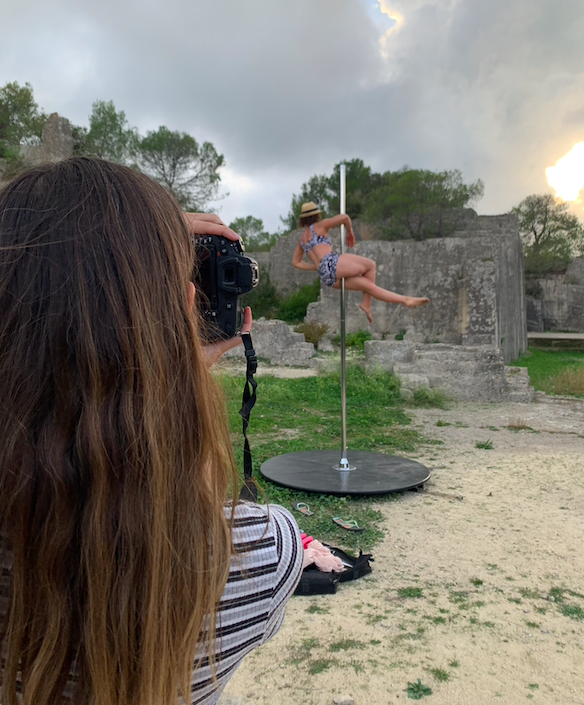 A woman taking a photo of a woman pole dancing outdoors on a circular platform with ruins and trees in the background.