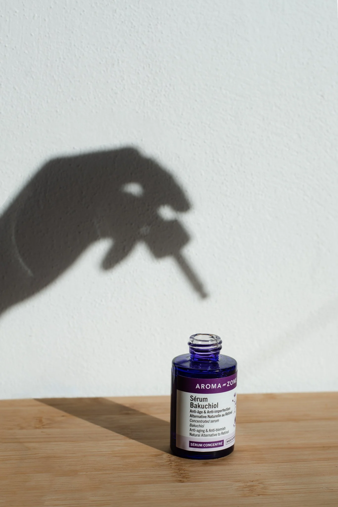 A blue glass bottle of skincare serum sitting on a wooden surface, with a shadow of a hand holding a dropper projected onto a white wall behind it.