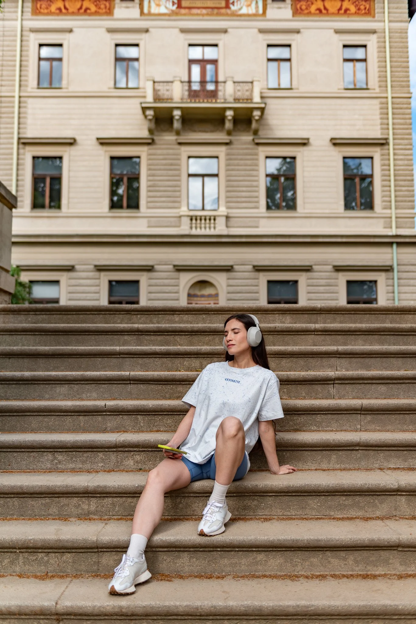 Young woman sitting on outdoor stone stairs, with a building in the background, wearing headphones, a light-colored t-shirt, shorts, and sneakers, holding a smartphone, with one eye closed and relaxed expression.