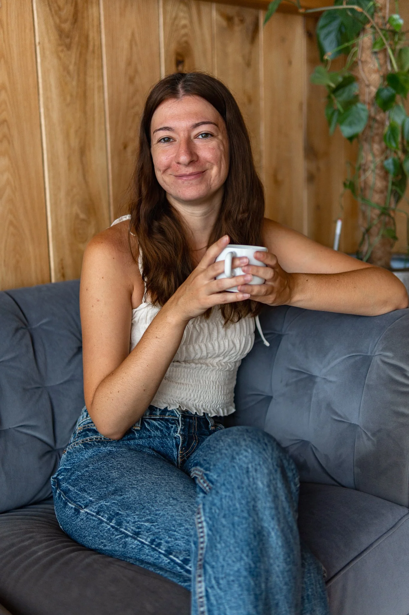 A woman with long brown hair sitting on a gray couch, holding a white mug, and smiling at the camera. She is wearing a white sleeveless top and blue jeans, with a wooden wall and green plant in the background.