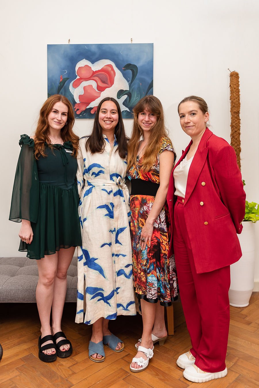 Four young women standing together indoors in front of a modern painting, smiling at the camera, dressed in fashionable outfits.