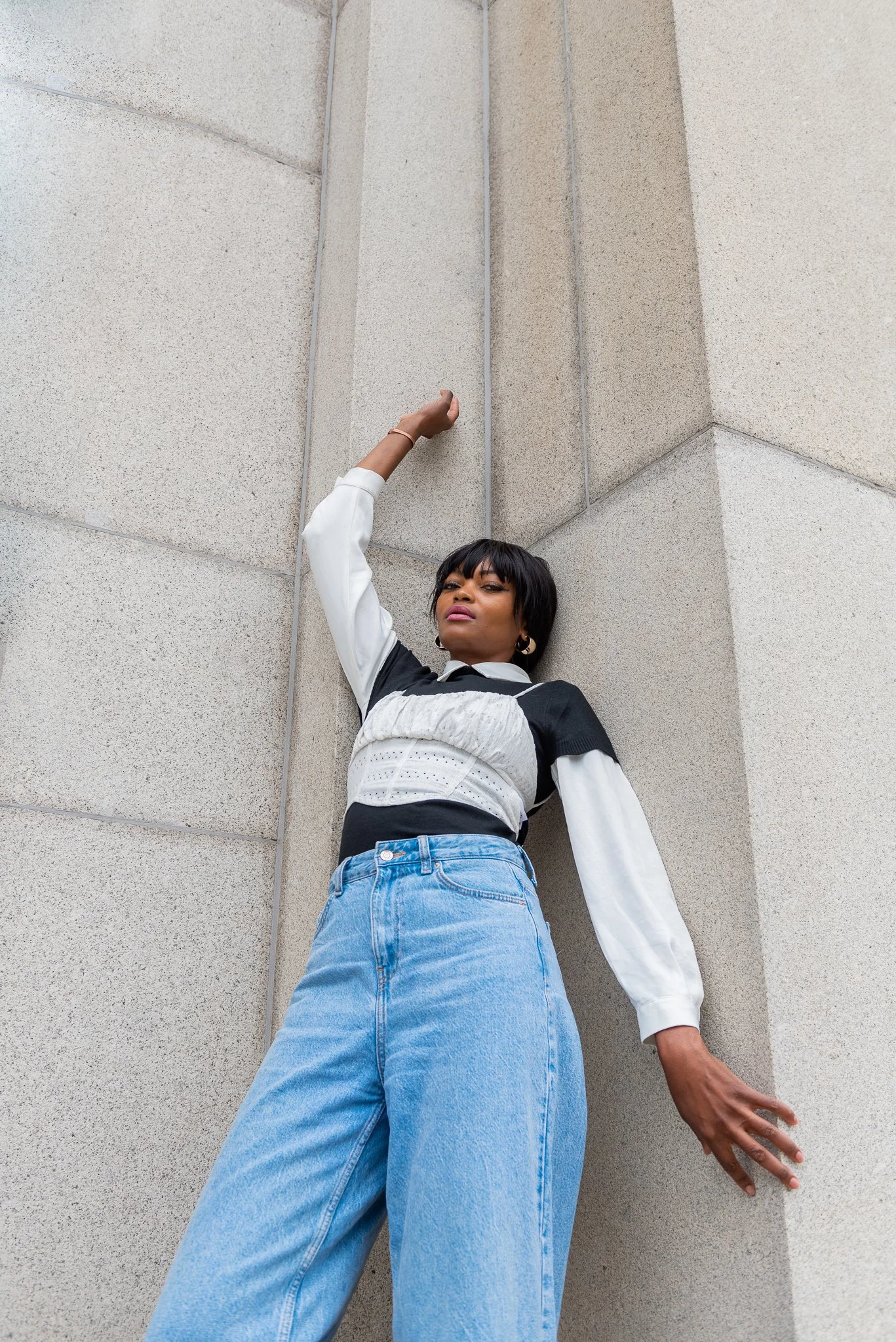 Young woman leaning against a building wall with her arm raised in a fashion pose, wearing a black and white top and high-waisted blue jeans.