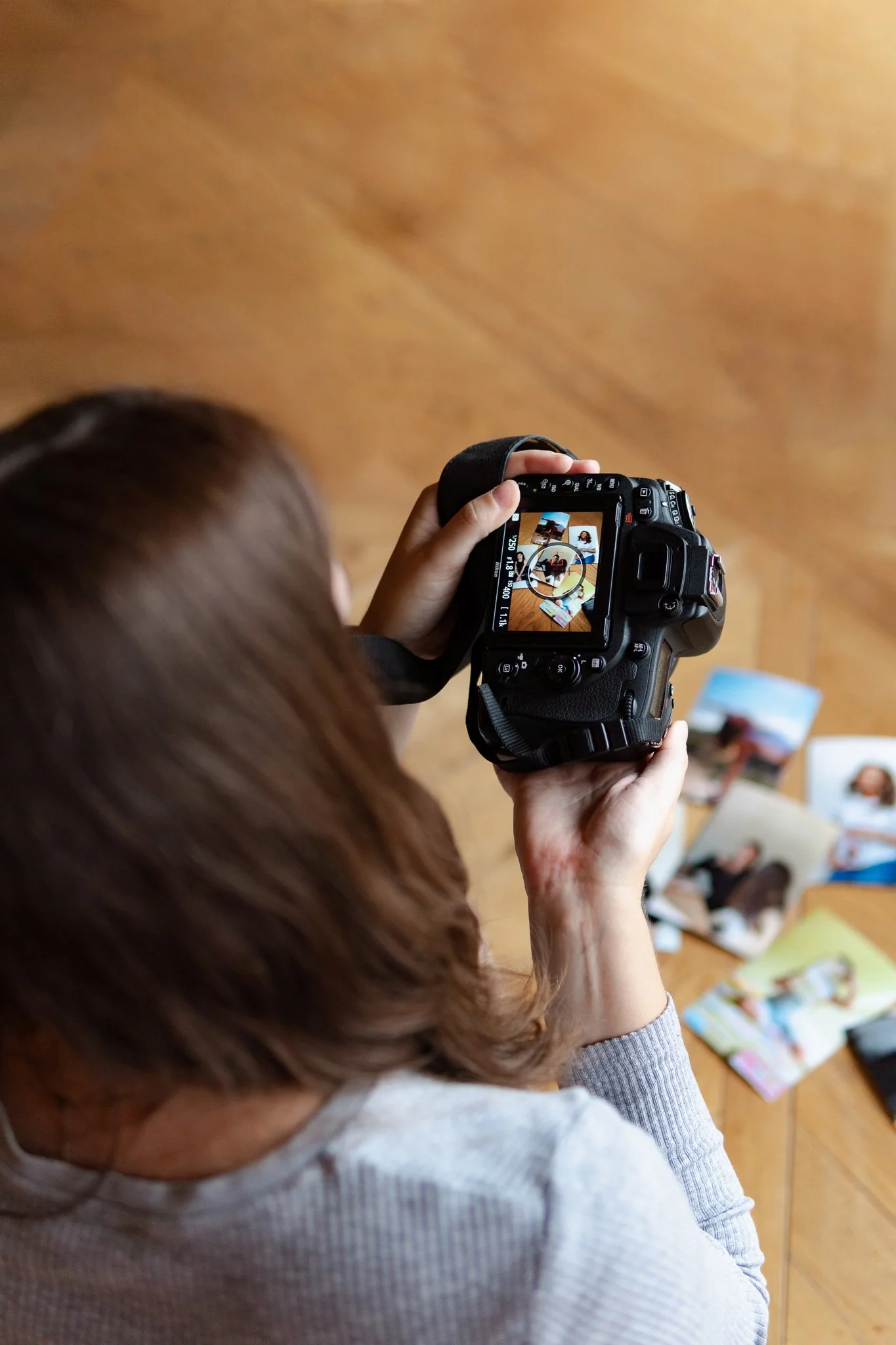 A woman with brown hair takes a photo of printed photographs on a wooden table with a digital camera.