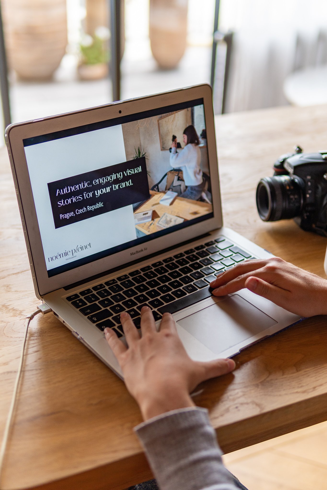 A person using a MacBook Air laptop on a wooden table with a camera nearby, displaying a presentation slide that reads 'Authentic, engaging visual stories for your brand' in a bright room with large windows.