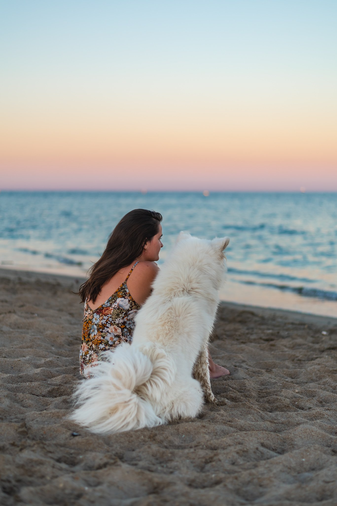 A woman and a large white dog sitting on a sandy beach during sunset, facing the ocean.