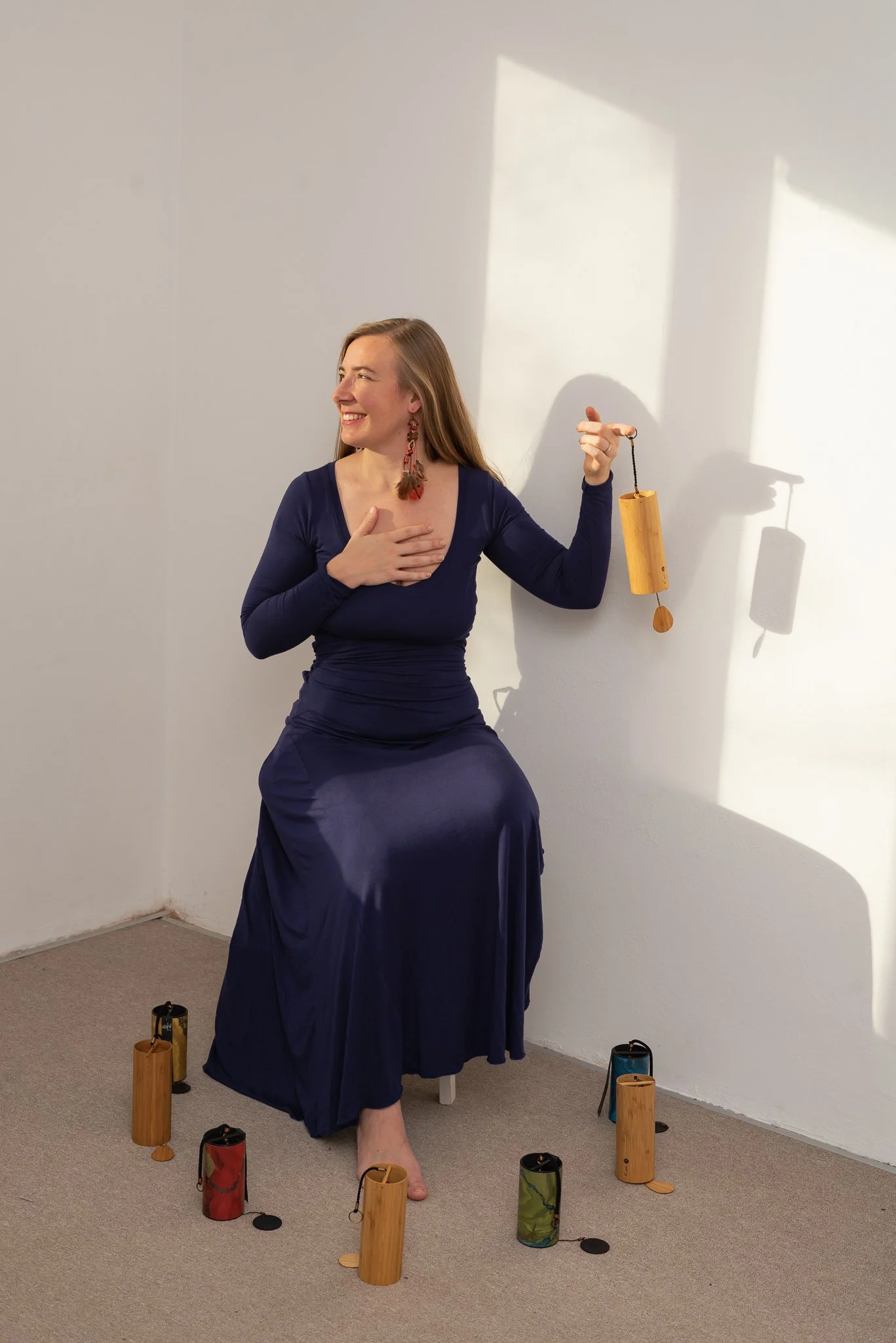 A woman in a long blue dress seated on a small stool in a room with plain white walls. She is smiling, touching her chest with her left hand, and holding a wooden percussion instrument (caxixi) with her right hand. The woman is barefoot, and there are six similar instruments placed on the floor around her, some in colorful cases.