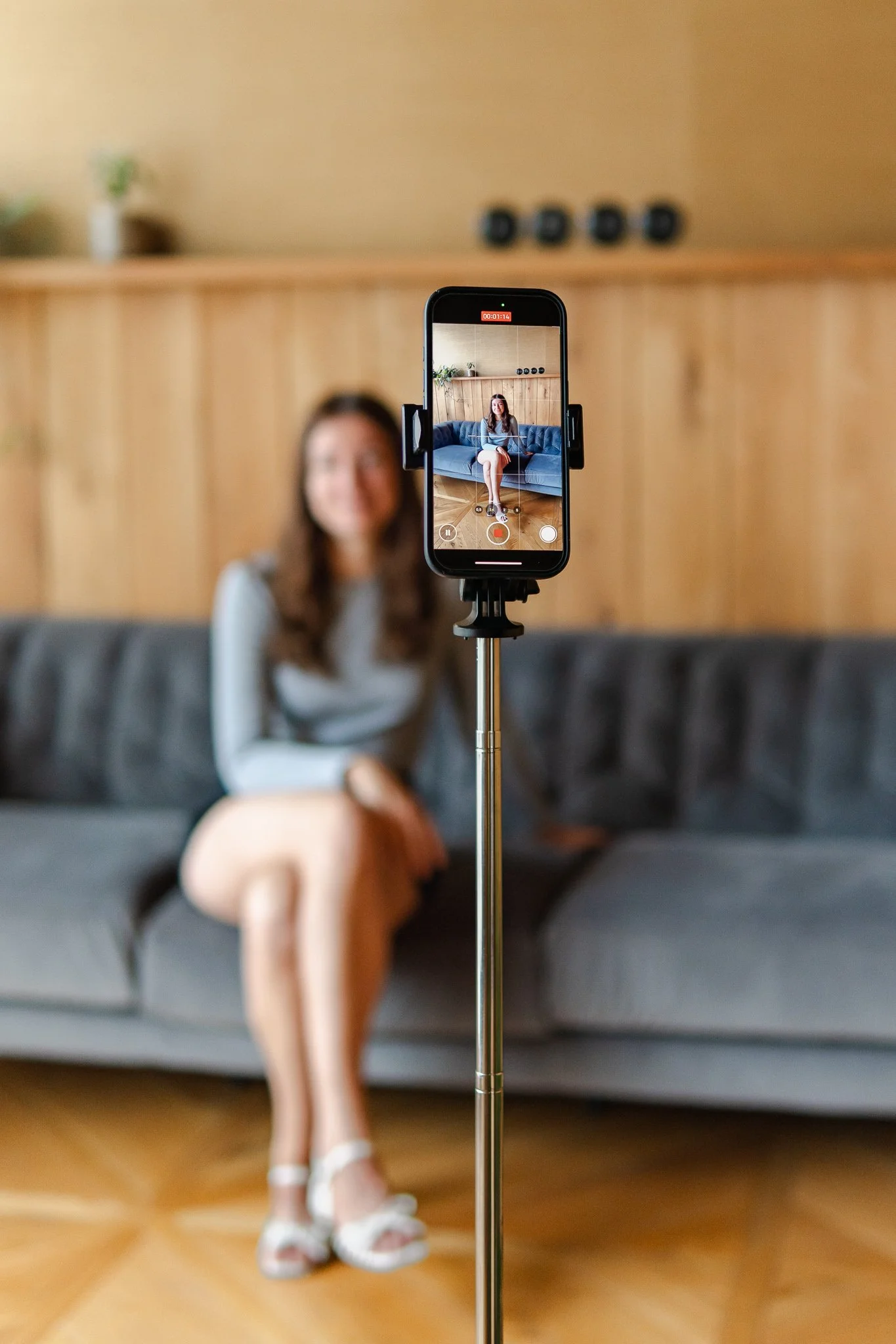 A woman sitting on a gray sofa with legs crossed, recorded on a smartphone mounted on a tripod, in a cozy room with wooden paneling and a light-colored wall.