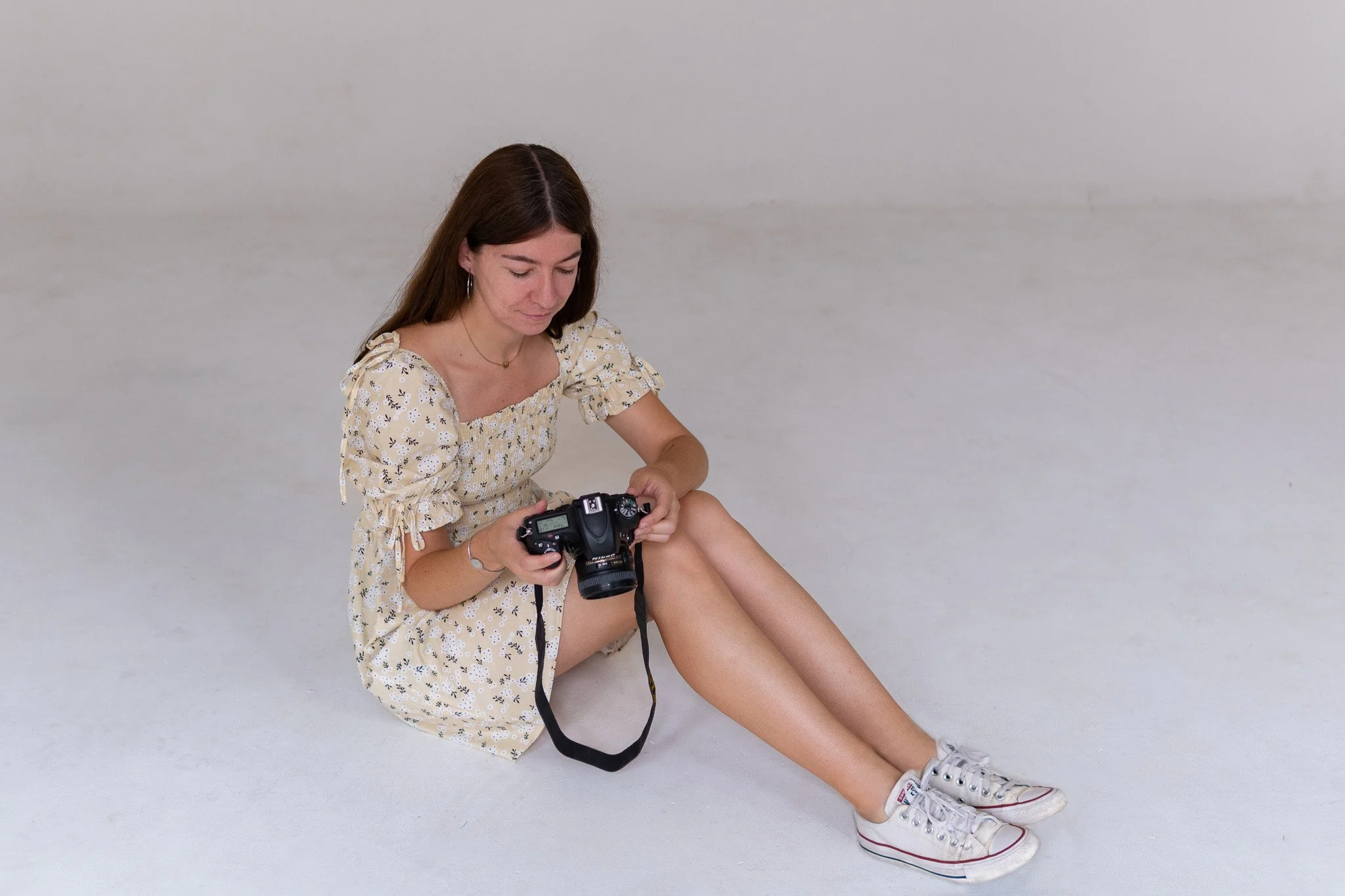 Young woman sitting on white floor, holding a DSLR camera and looking at its screen, wearing a floral dress and white sneakers.