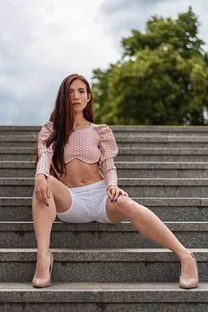 A woman sitting on outdoor steps in a pink cropped top, white shorts, and beige high heels, with greenery and trees in the background. Fashion photography in Prague.