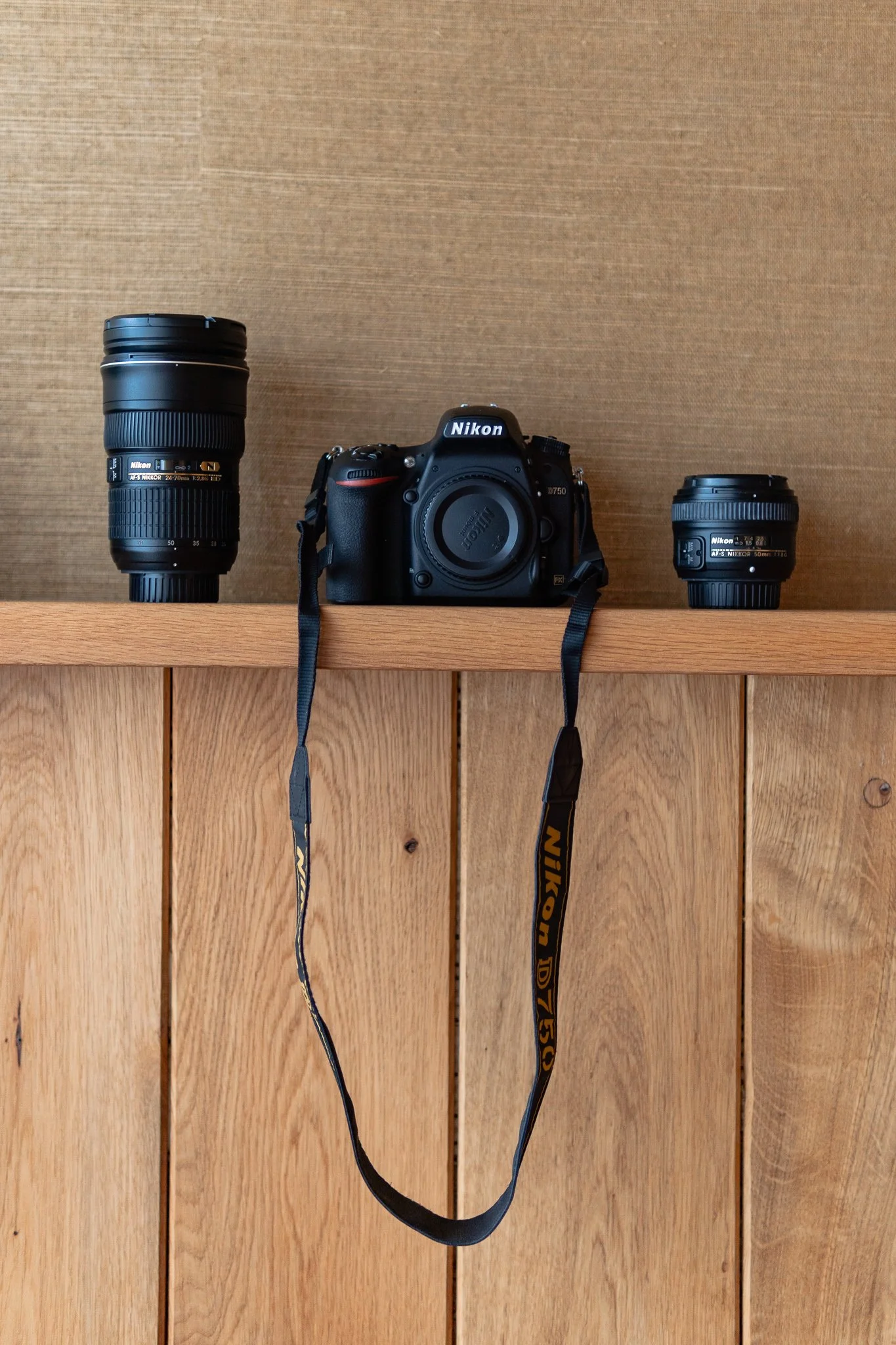A Nikon DSLR camera with two Nikon lenses on a wooden shelf against a brown wall.