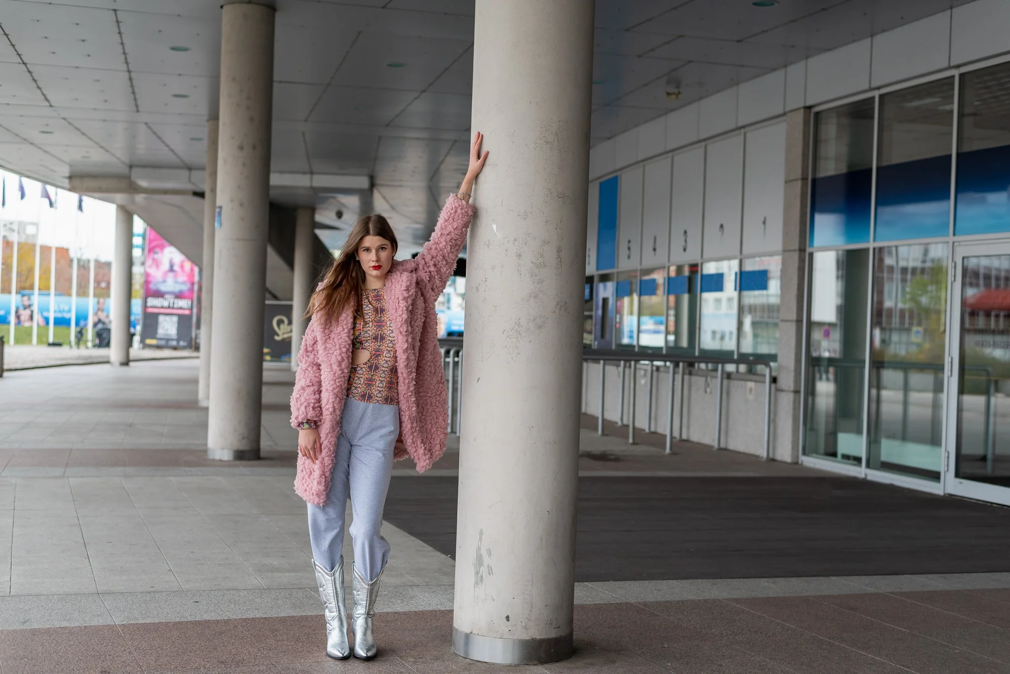 Young woman in pink fuzzy coat and cowboy boots leaning against a large white column outside a modern building.