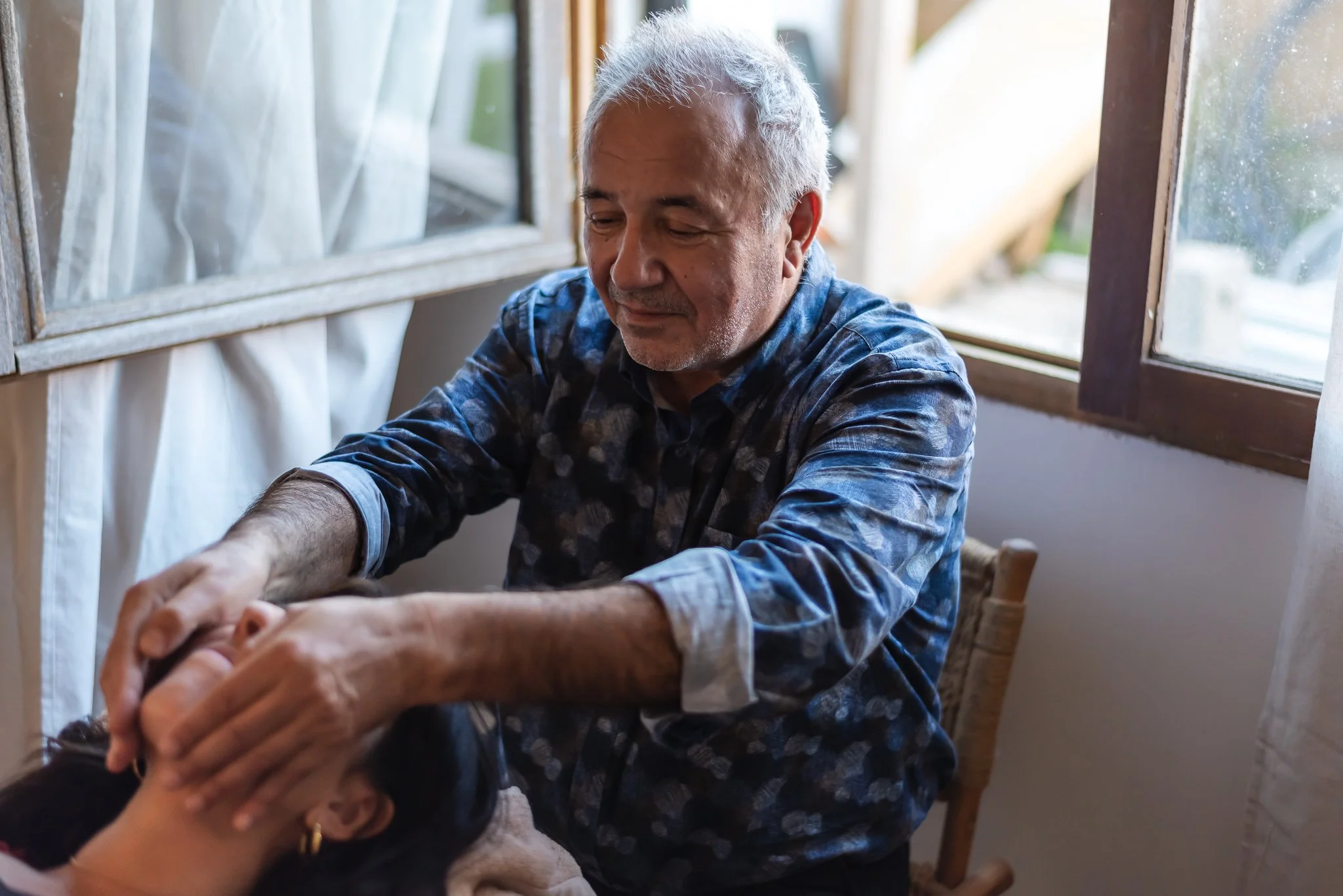 An elderly man in a blue patterned shirt giving a gentle facial massage to a woman with dark hair, inside a room with sunlight coming through open windows.
