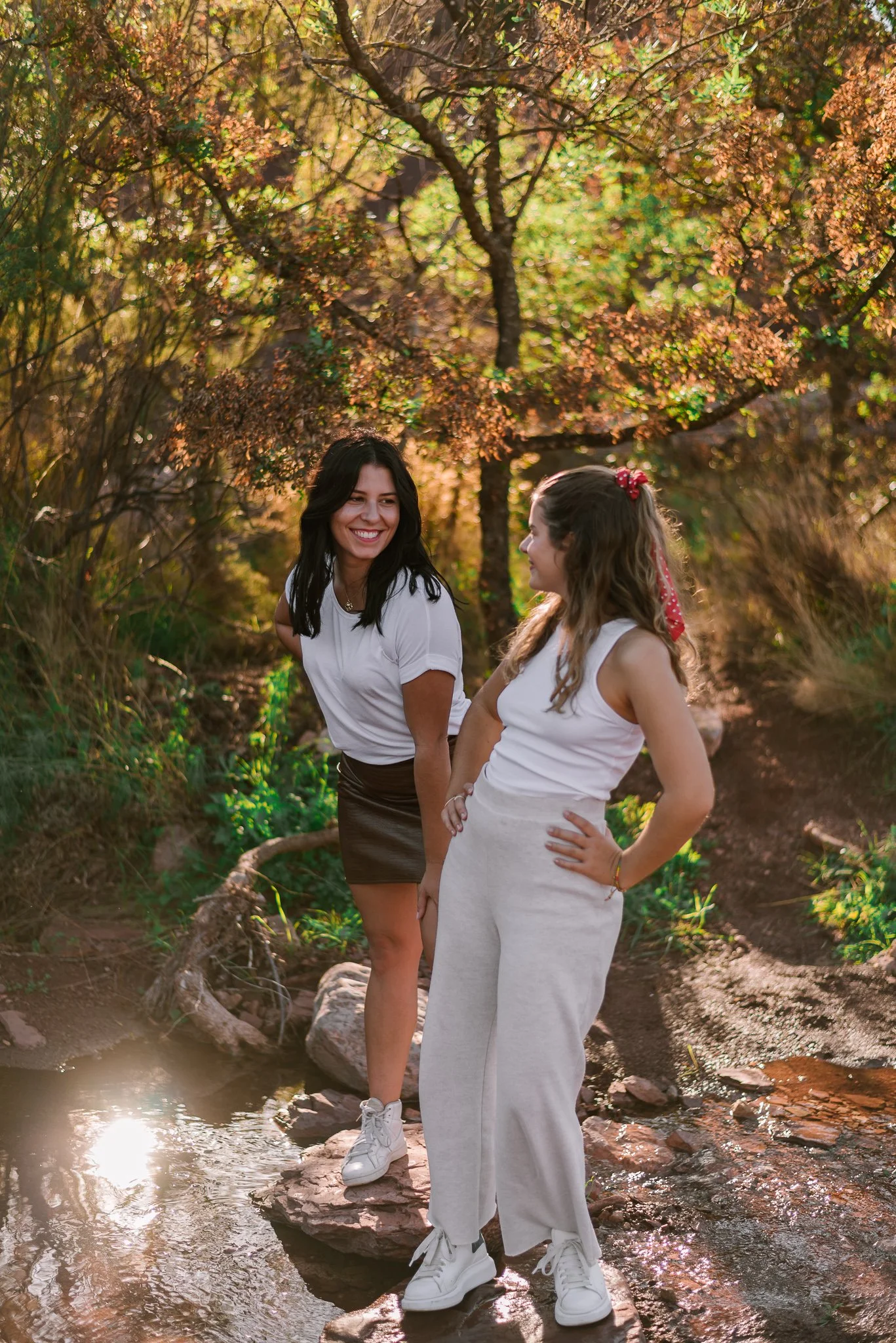 Two young women smiling and laughing on rocks by a small creek in a forest during sunset.