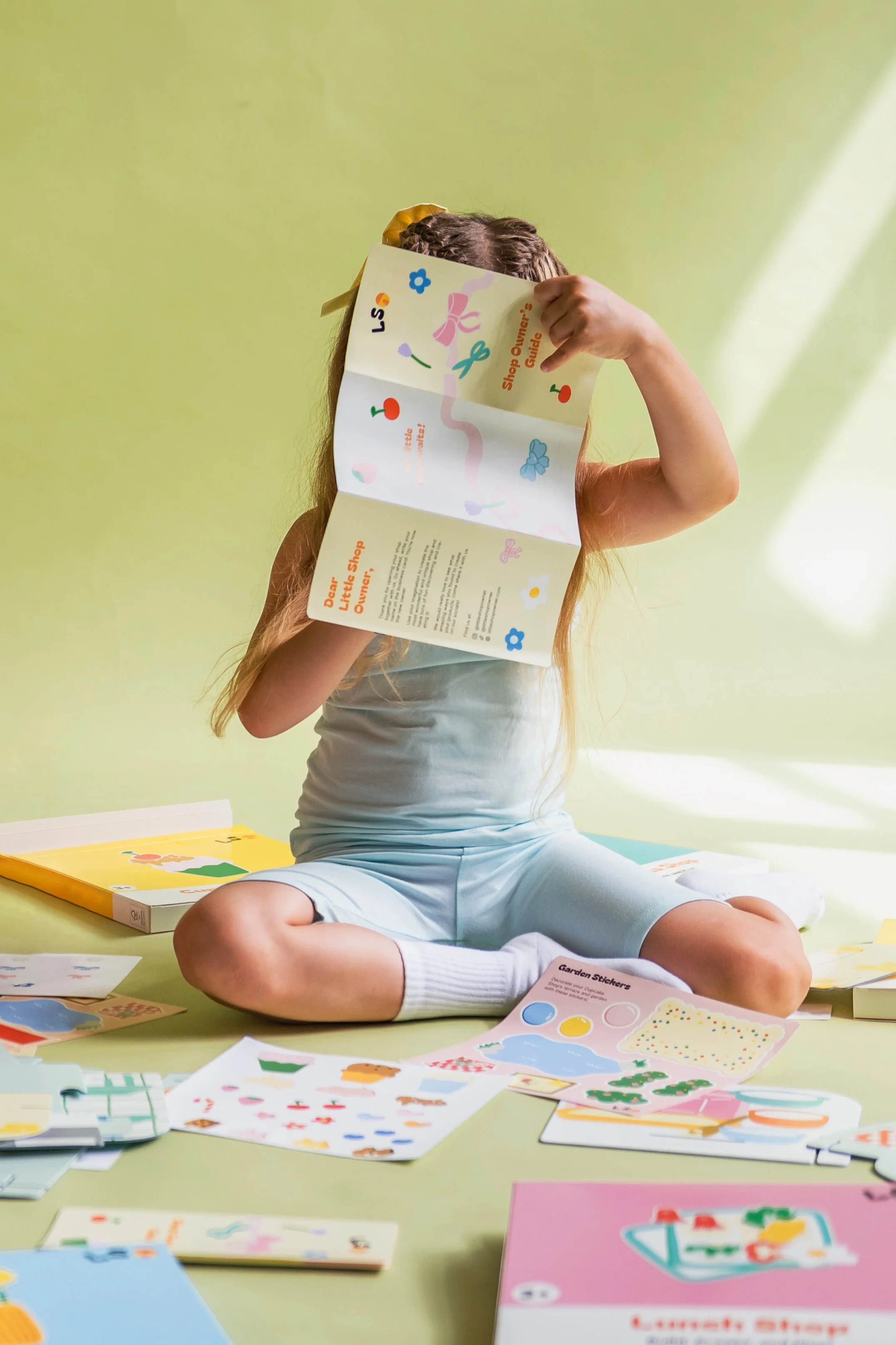 Child sitting cross-legged on a light green surface, holding a colorful paper, surrounded by various sheets of paper and books with colorful illustrations.