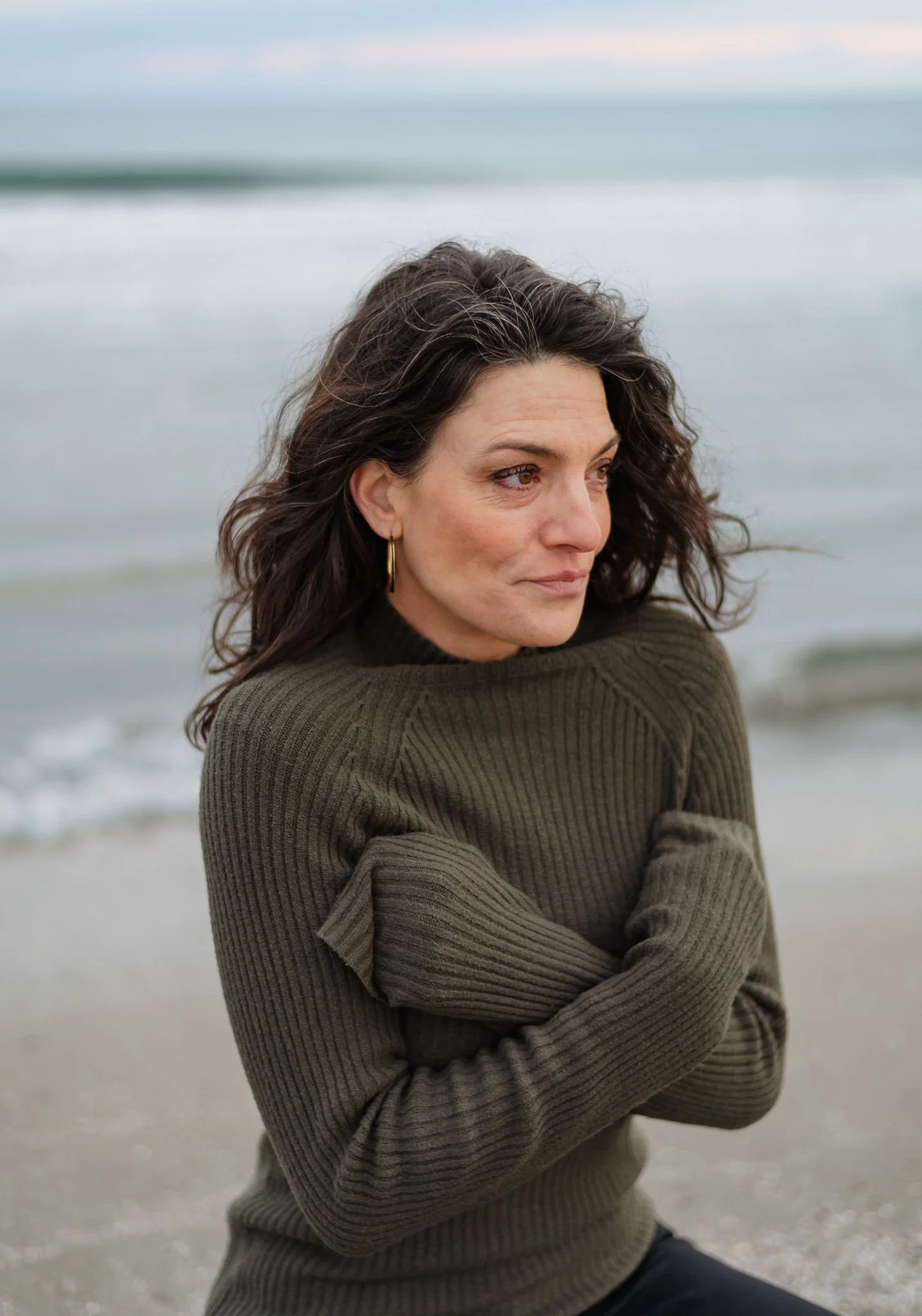 A woman with curly brown hair and gold hoop earrings standing on a beach near the ocean, wearing a dark green, ribbed sweater with crossed arms, looking contemplative.