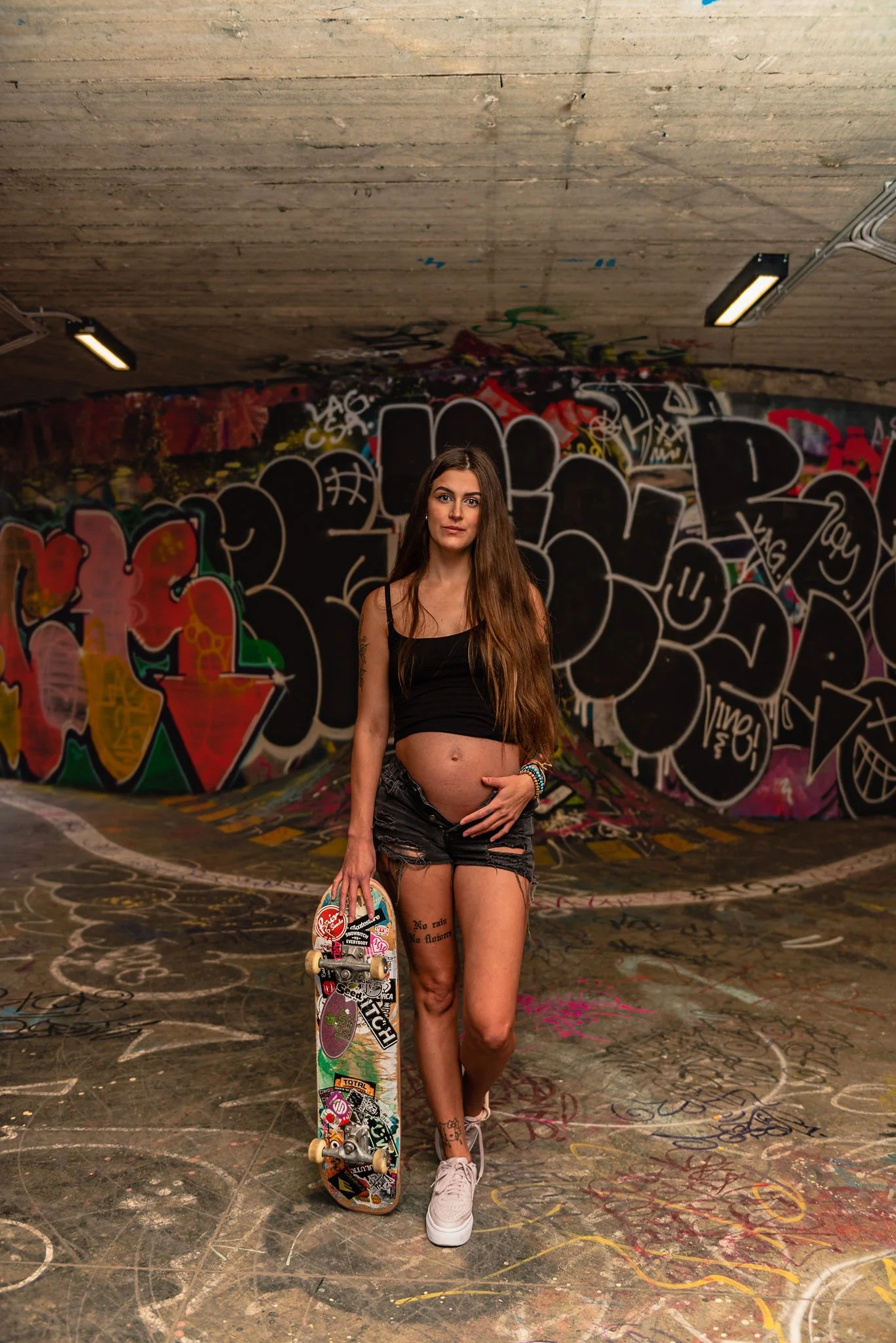 A pregnant woman holding a skateboard in an underground skate park with colorful graffiti on the walls in Holešovice, Prague.