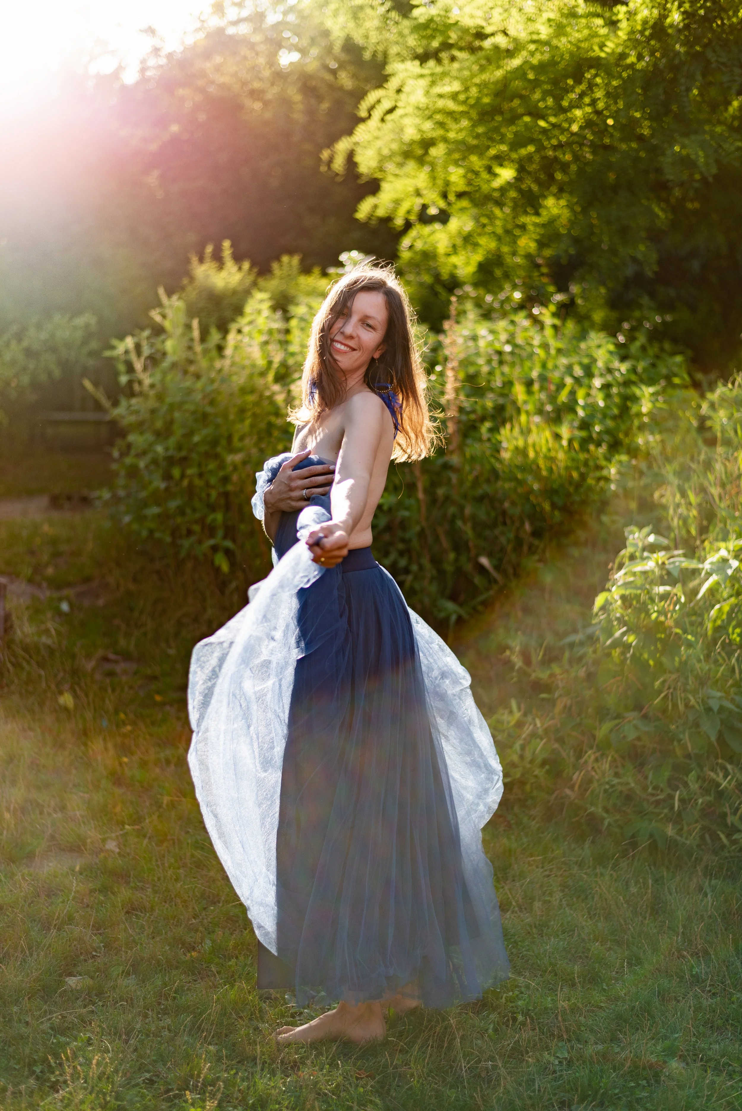 A woman with long brown hair, smiling and holding a cloth in a natural outdoor setting with green trees and sunlight in the background.