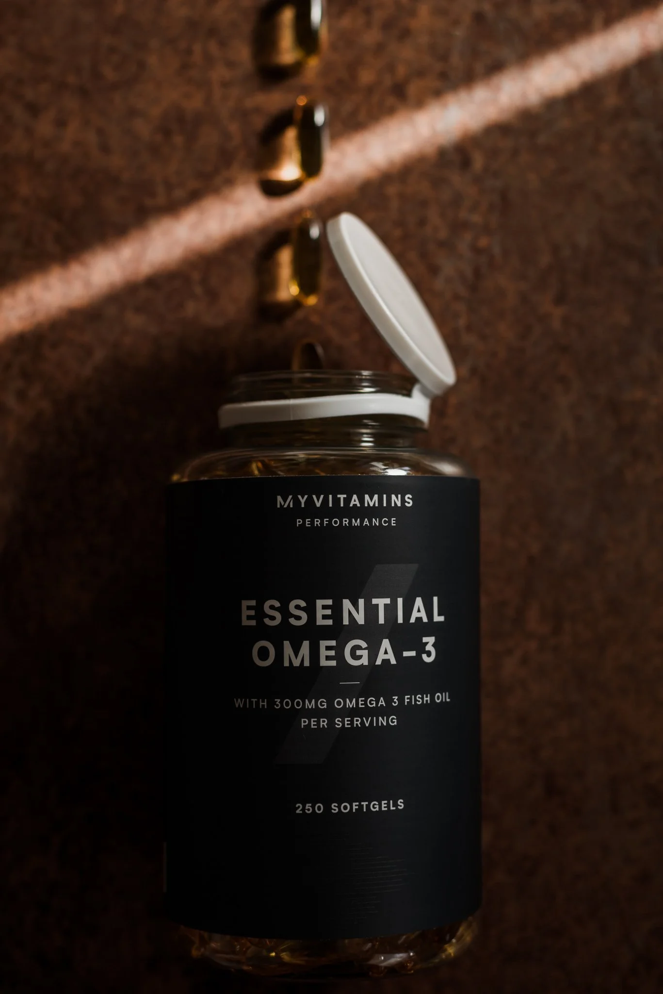 A bottle of MYVITAMINS Performance Essential Omega-3 softgels with an open lid, set against a textured brown background with metallic hooks and a beam of light casting shadows.