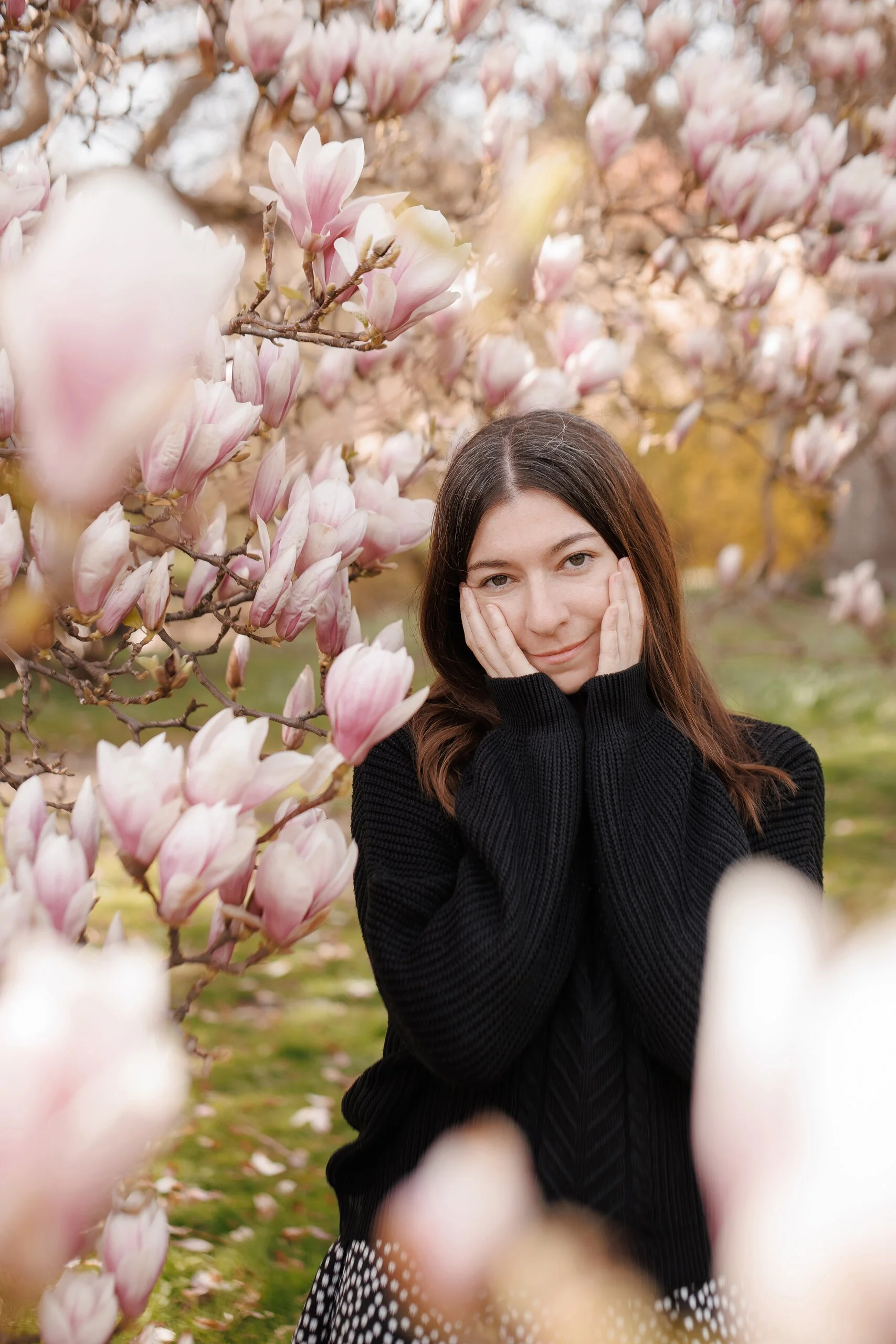 A woman with brown hair wearing a black sweater and black and white polka dot skirt, posing amidst blooming magnolia trees with pink and white flowers, covering her ears with her hands and smiling.