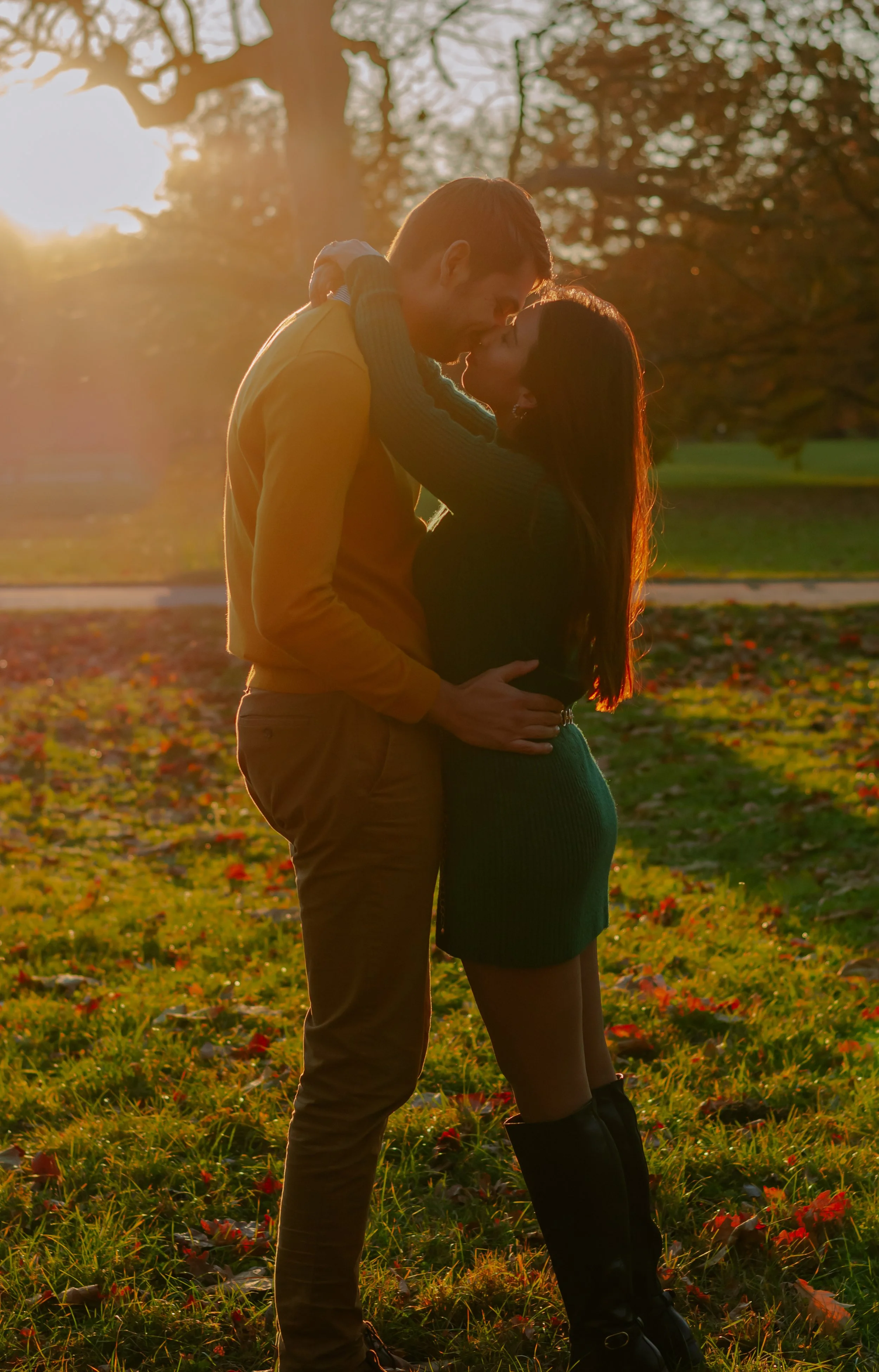 A couple embracing and about to kiss outdoors during sunset, with trees and autumn leaves in the background.