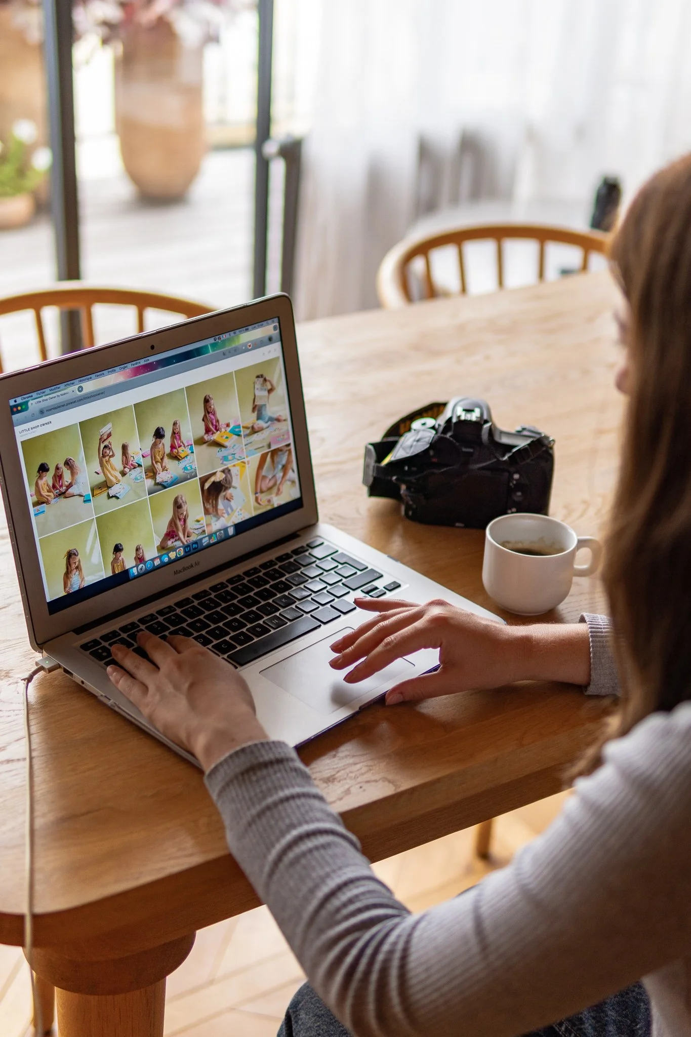 A woman sitting at a wooden table using a MacBook to view a photo gallery of kids playing with toys.