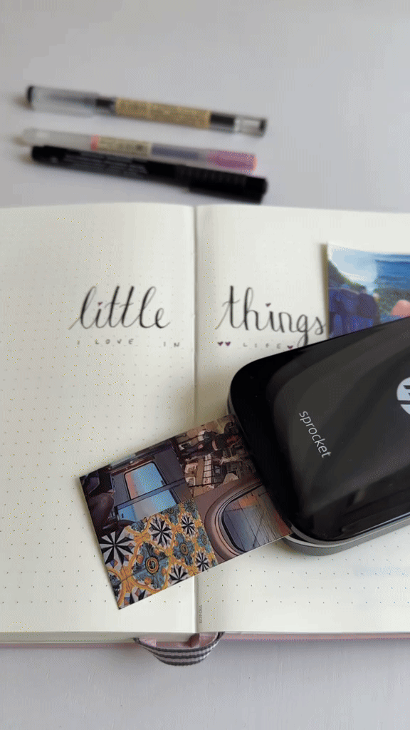 Open notebook with handwritten words 'little things' and a small photo of a colorful patterned room, with a black HP sprocket photo printer on top.