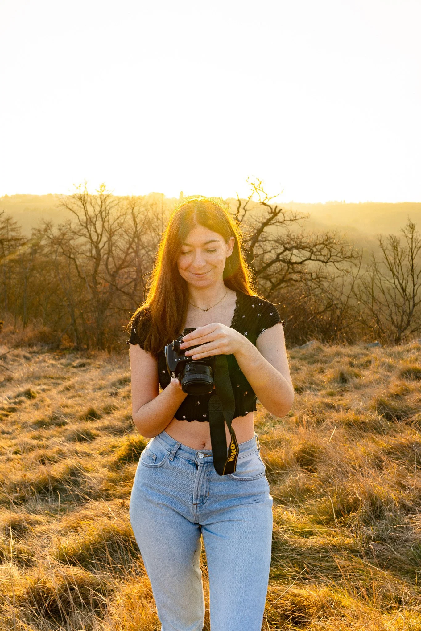 A young woman with long auburn hair wearing a black cropped top and light blue jeans stands in a grassy field at sunset, holding a DSLR camera and smiling softly.