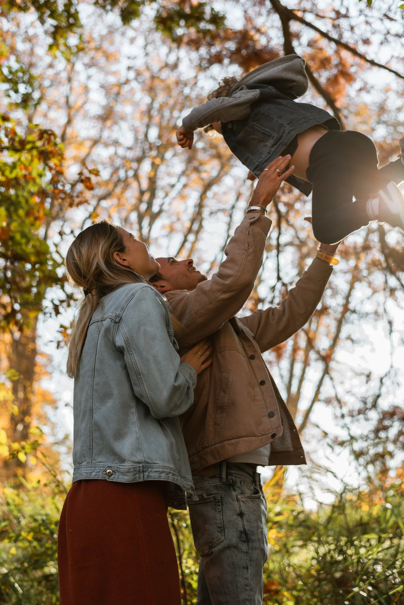 A family outdoors in autumn, with trees and fall foliage in the background. A man is lifting a young girl into the air while a woman looks on affectionately. Family photoshoot in Prague.