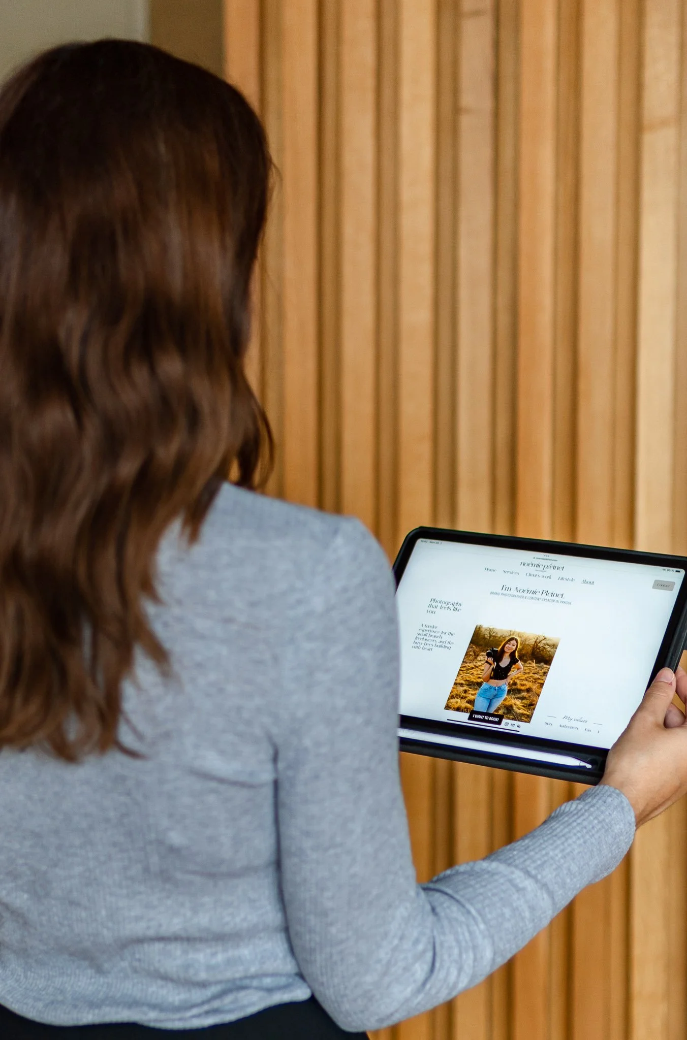 Back view of a woman with brown wavy hair reading an e-book on a tablet, sitting against a wooden panel background.
