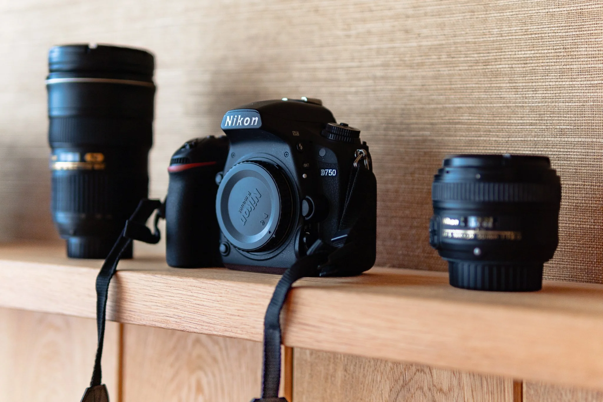 A Nikon DSLR camera with two camera lenses placed on a wooden shelf against a beige textured wall.