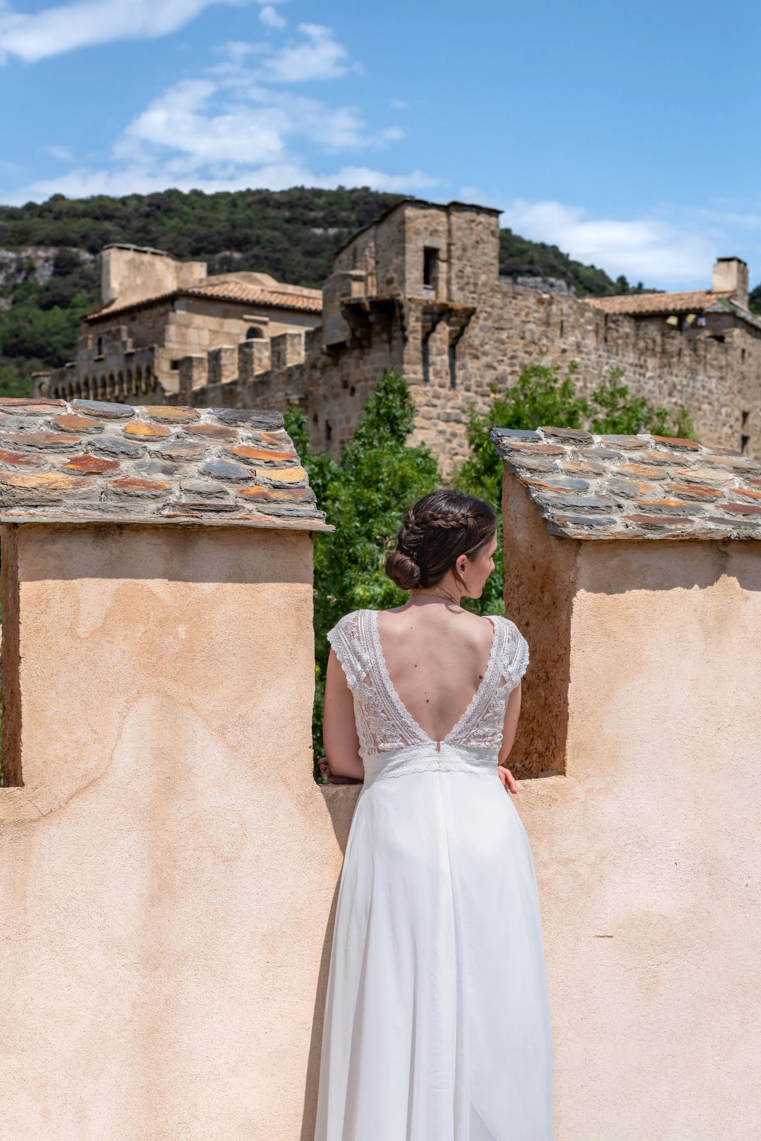 A woman in a white lace dress with an open back, standing on a terrace and looking toward an ancient stone castle on a hill in the background, with a blue sky and some clouds.