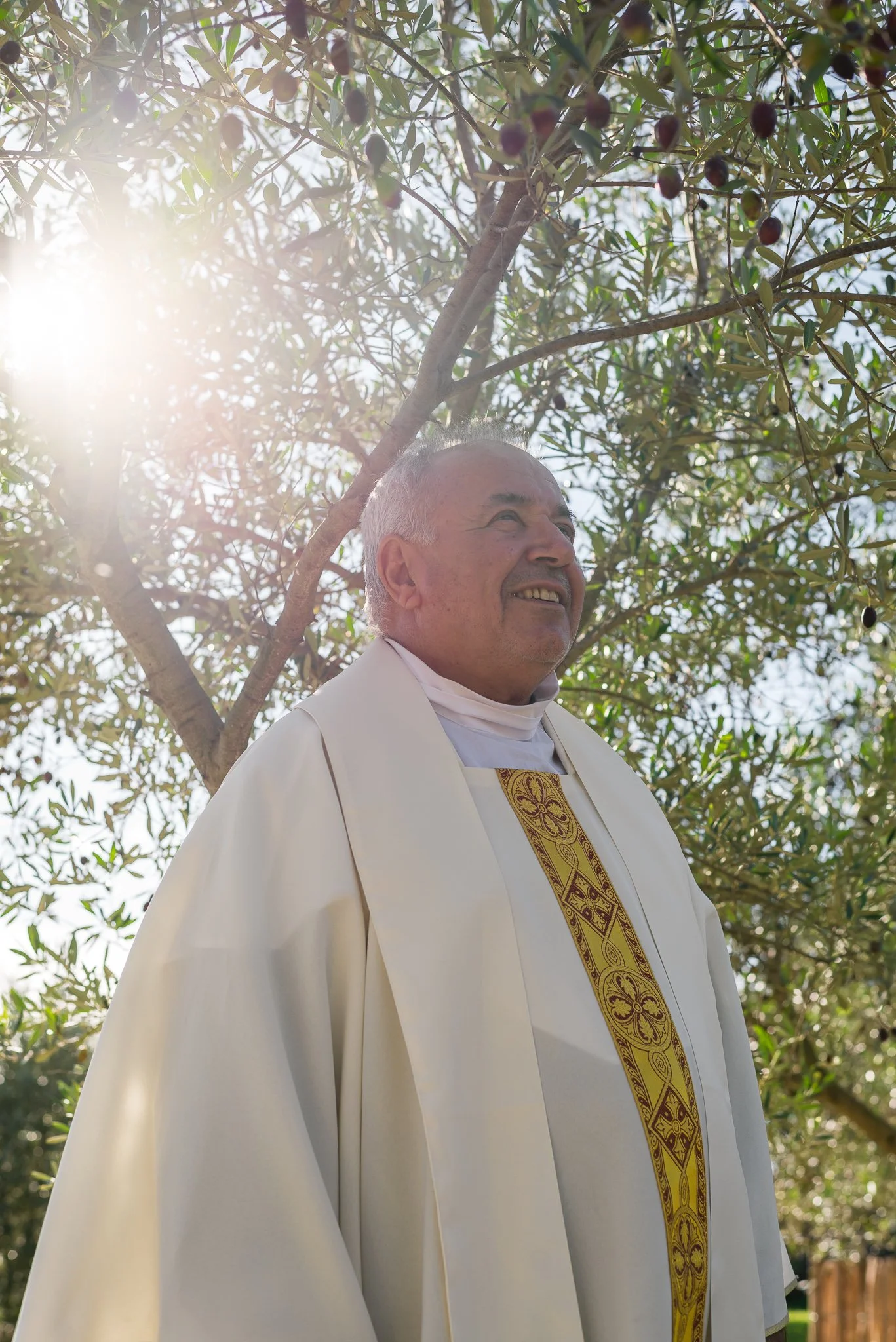 Clergy member wearing white robes with gold embroidery, standing outdoors under a tree with sunlight shining through the leaves.