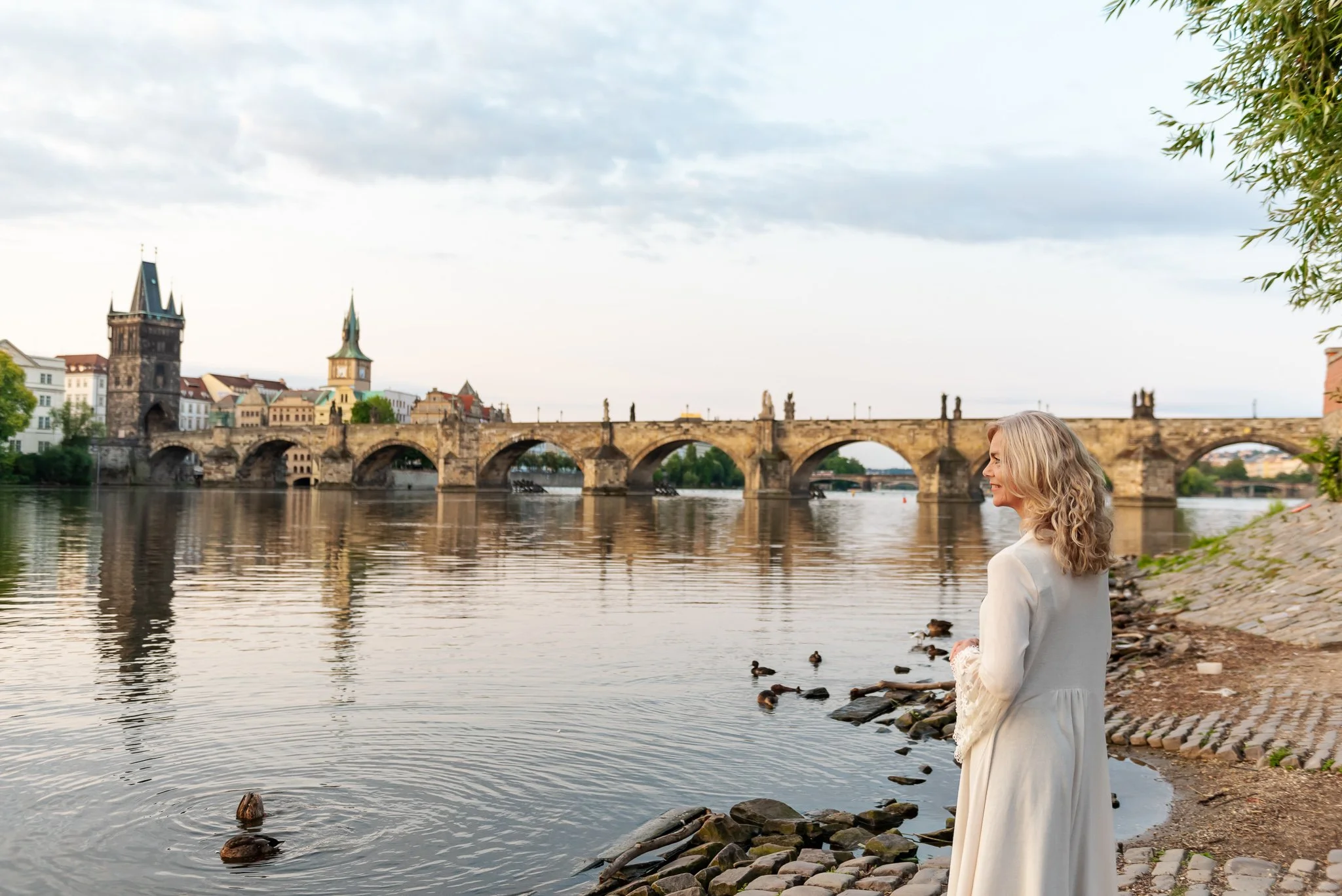A woman with curly blonde hair smiling by the riverbank with ducks, with historic buildings and a stone bridge in the background.