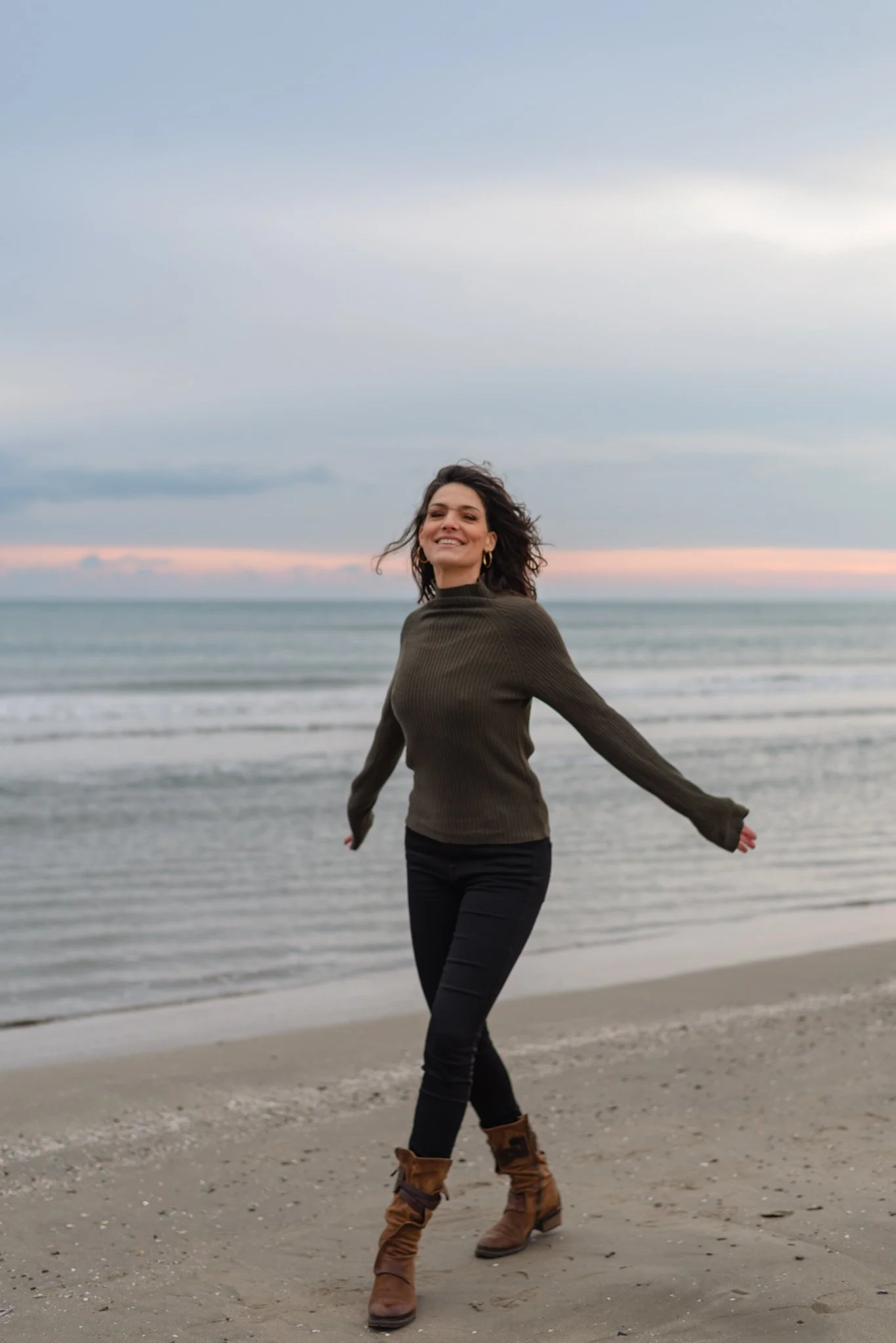 A smiling woman with curly brown hair wearing a green sweater, black pants, and brown boots, standing on a beach with the ocean and a cloudy sky in the background.