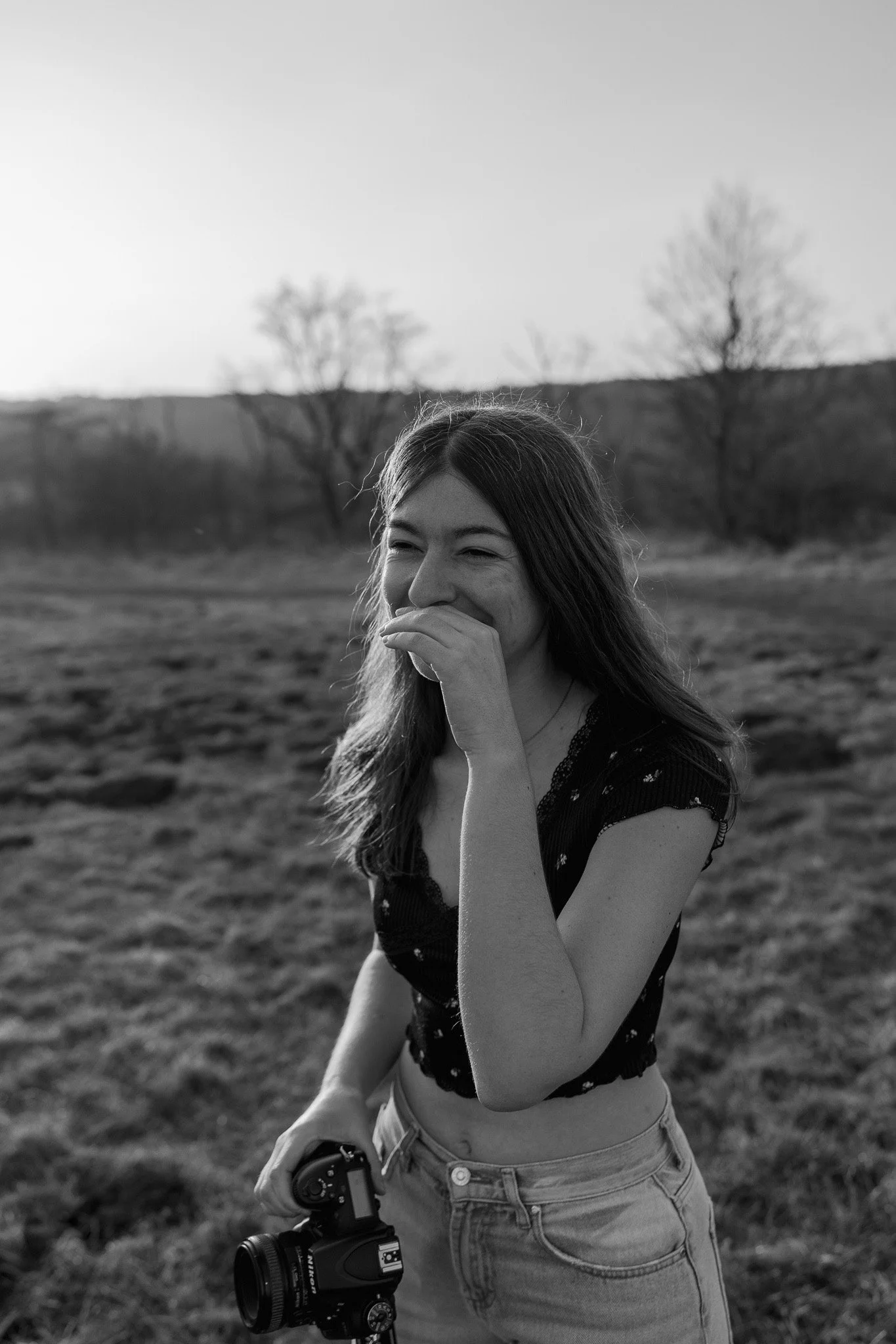 A young woman standing outdoors in a field, holding a camera, smiling and laughing, with trees and a hill in the background, black and white photo.