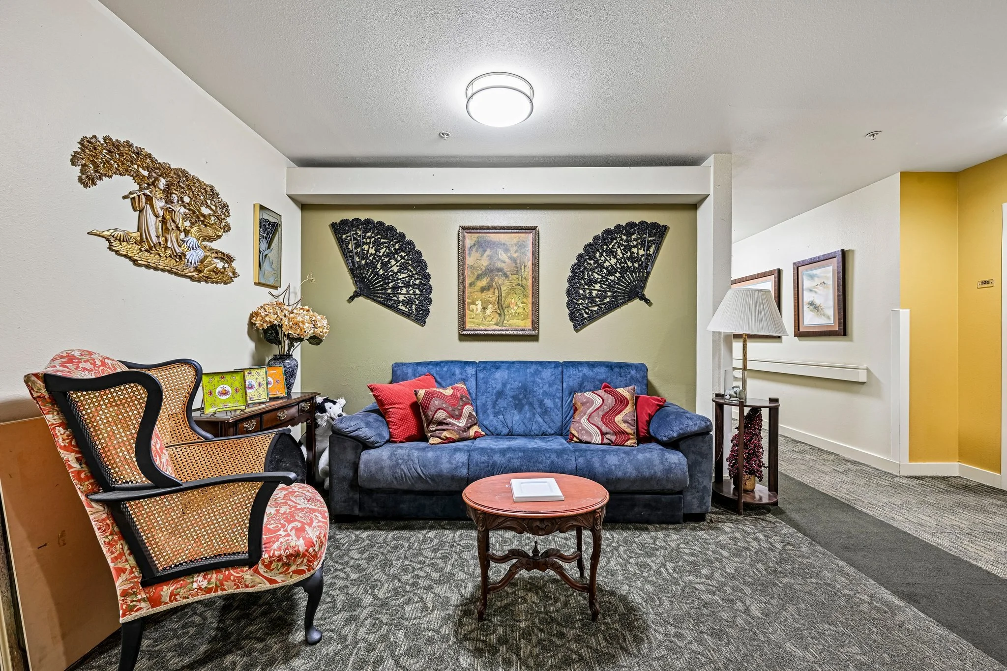 Interior of a living room with a blue sofa, colorful pillows, a round wooden coffee table, a floral patterned armchair, wall decorations including black fans and framed pictures, a yellow accent wall, and various decorative items like a lamp and flowers.