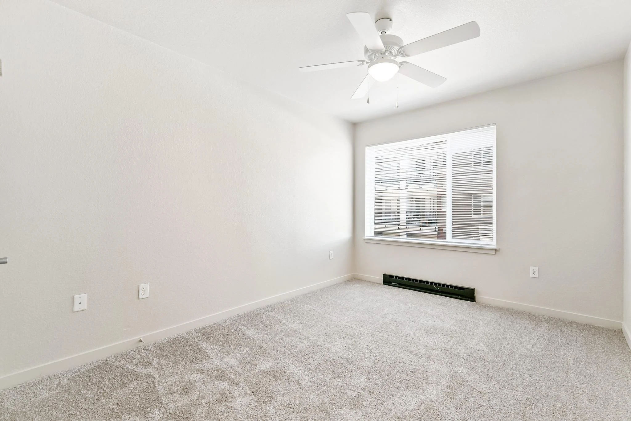 Empty room with beige carpet, white walls, a large window with blinds, ceiling fan with light, and electrical outlets.