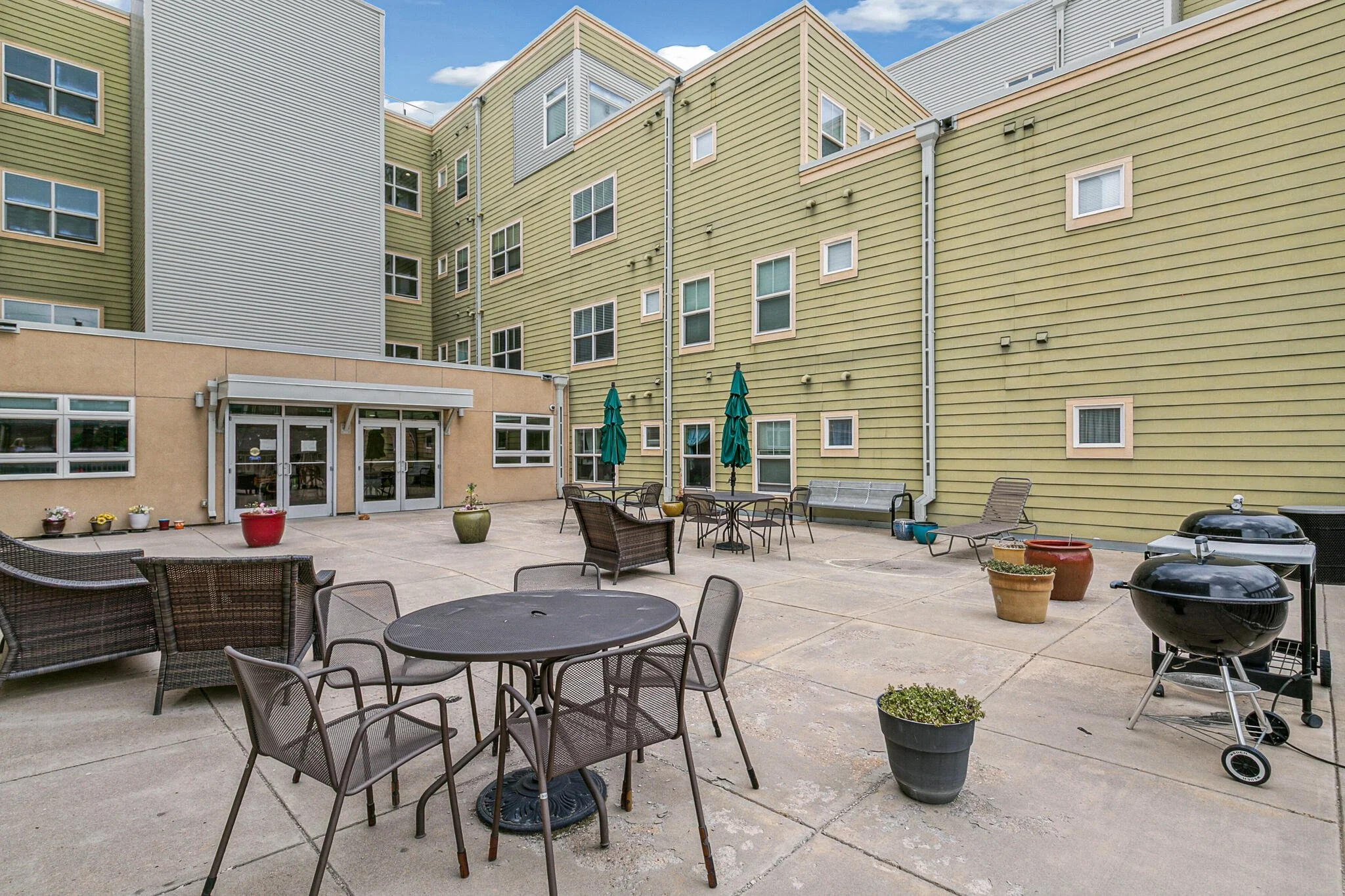 Apartment building courtyard with outdoor tables, chairs, umbrellas, and potted plants, with a clear blue sky.