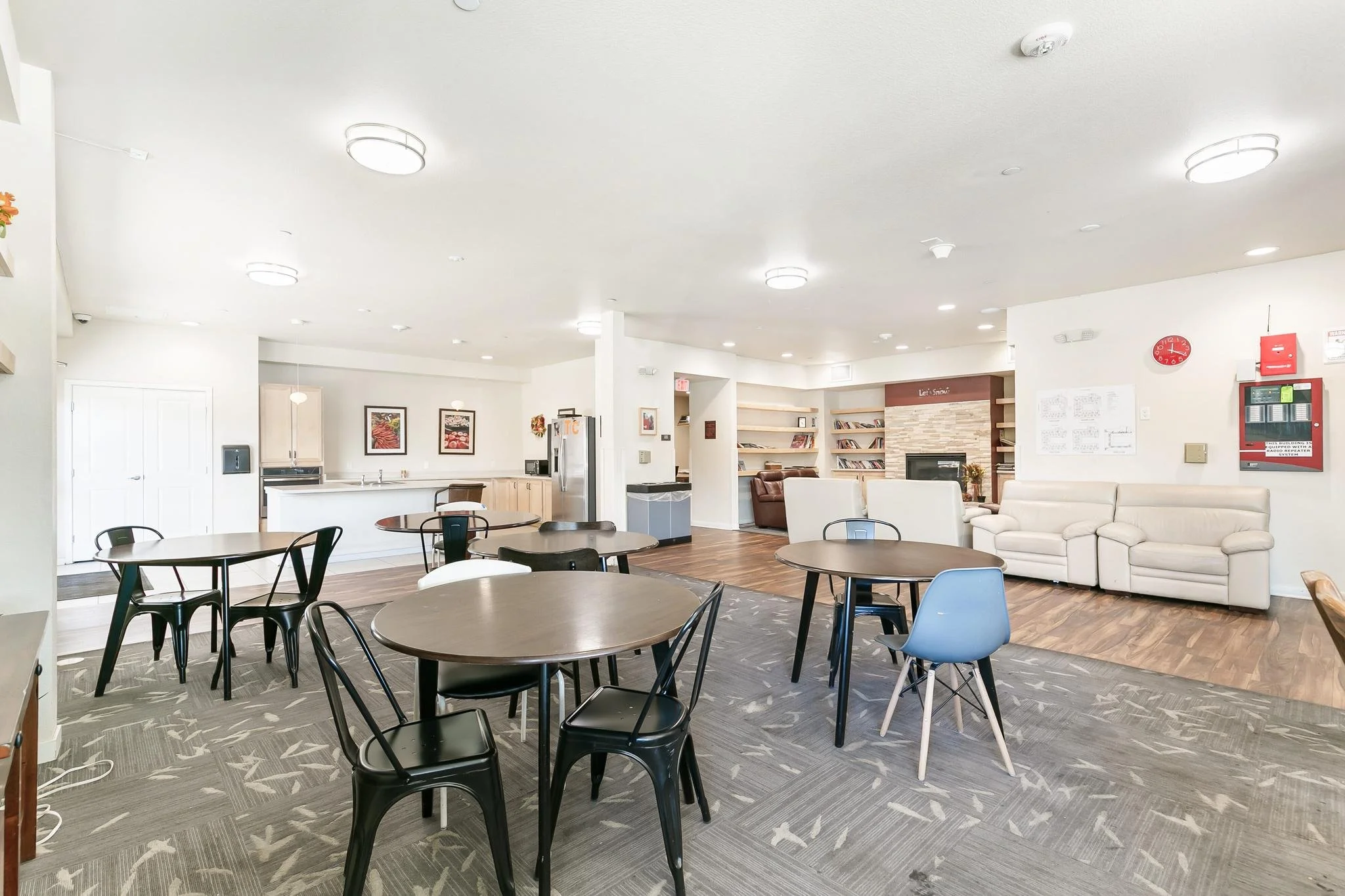 A spacious, modern communal area with multiple round tables and chairs, a white couch, a kitchenette with artwork, and a fireplace with bookshelves, well-lit with ceiling lights.