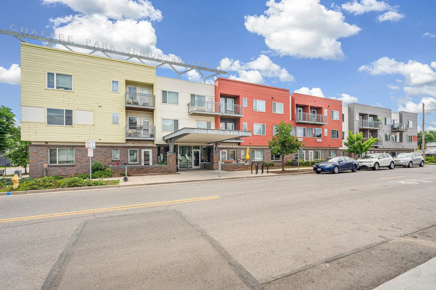 Front view of a multi-story apartment building with colorful exterior panels in yellow, white, red, and gray, located at 4580 North Tejon Street, Colorado Springs, Colorado, under a partly cloudy sky with parked cars and small trees in front.