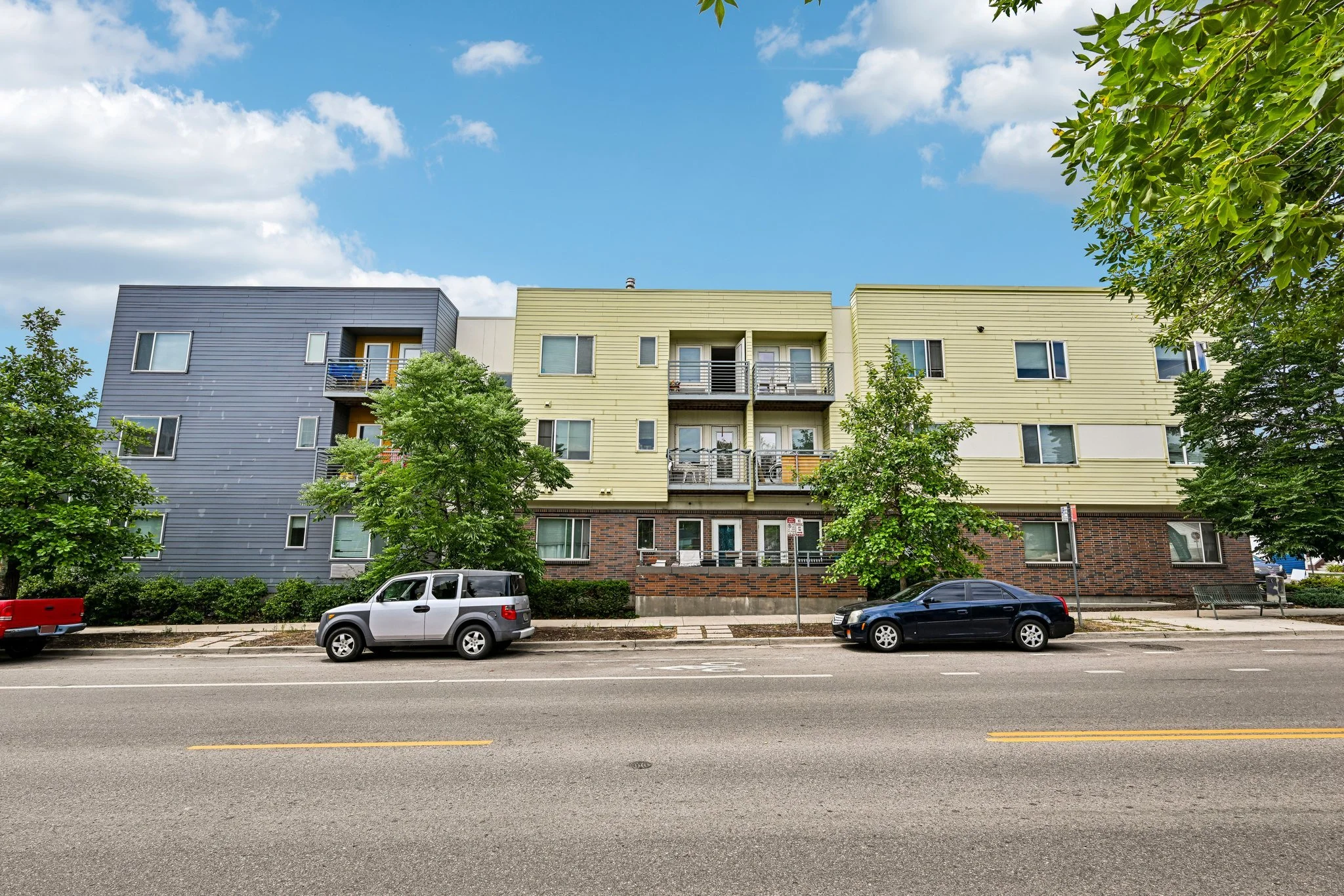 A modern multi-story apartment building with colorful paneling in blue, yellow, and beige, green trees in front, parked cars along the street, and a blue sky with clouds overhead.