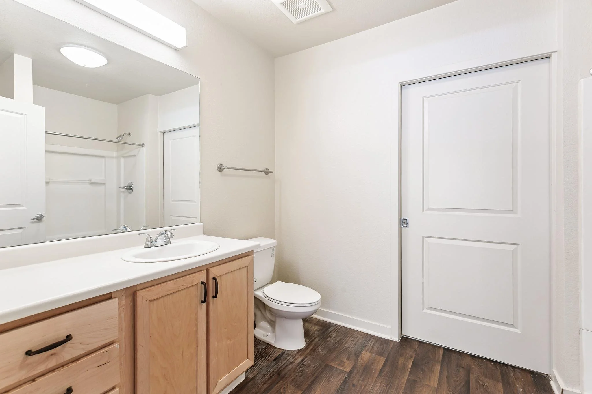 A small bathroom featuring a sink with a wooden cabinet, a mirror, a toilet, and a shower area with a glass door, white walls, dark wood flooring, and a wall-mounted towel bar.