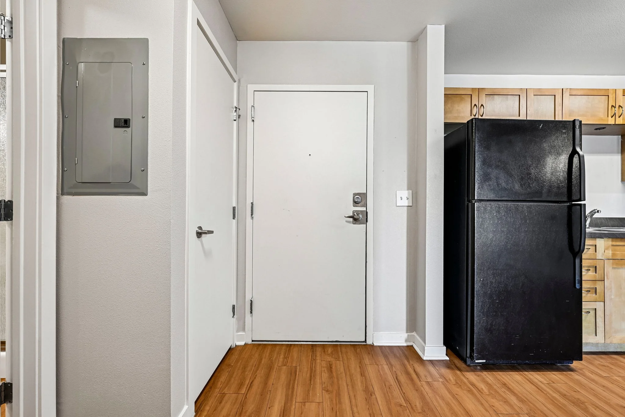 Interior view of a room with a white door, a black refrigerator, wooden cabinets, and hardwood flooring.