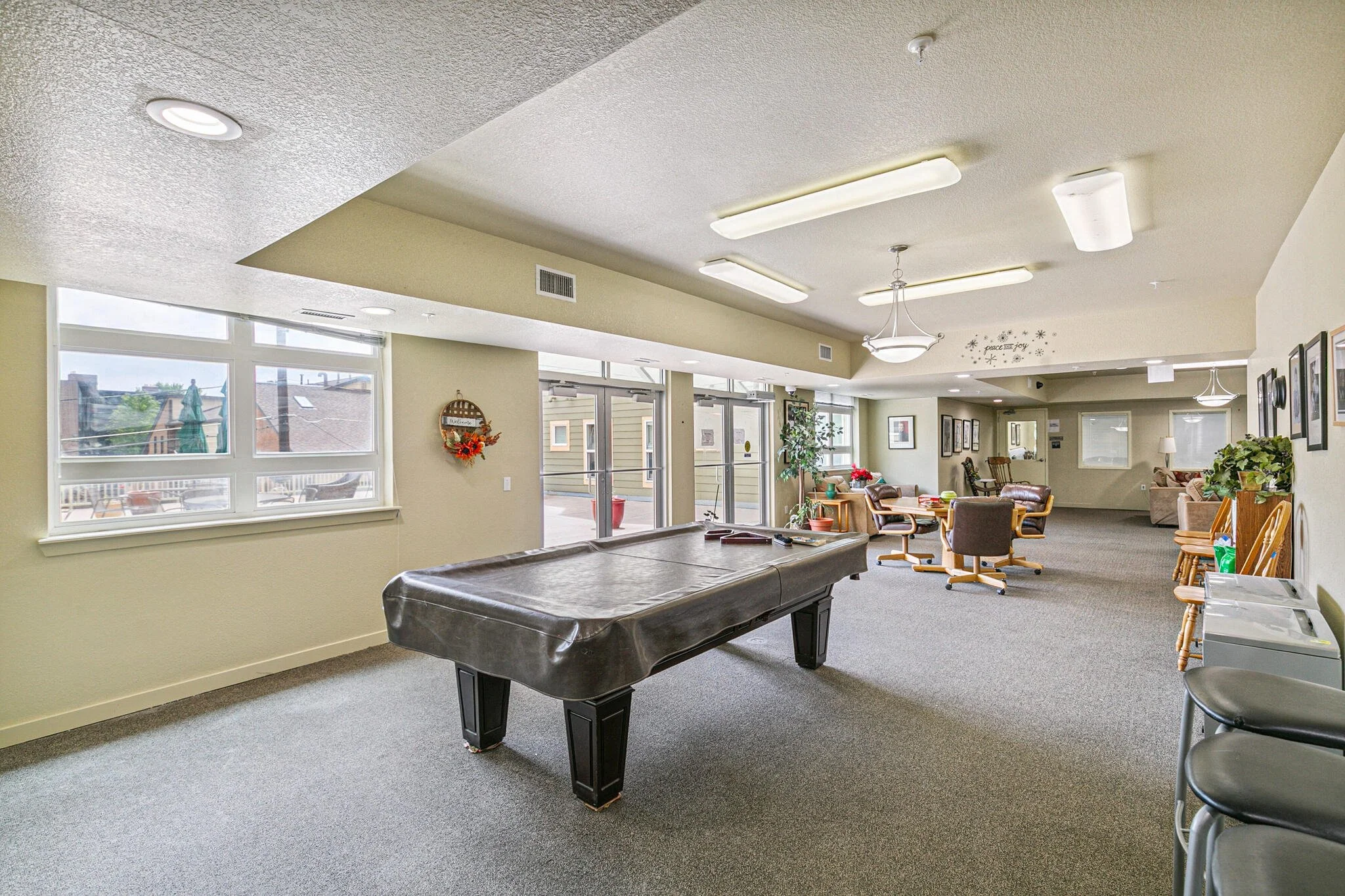 A spacious common room with large windows, chairs, a pool table, and wall decorations.