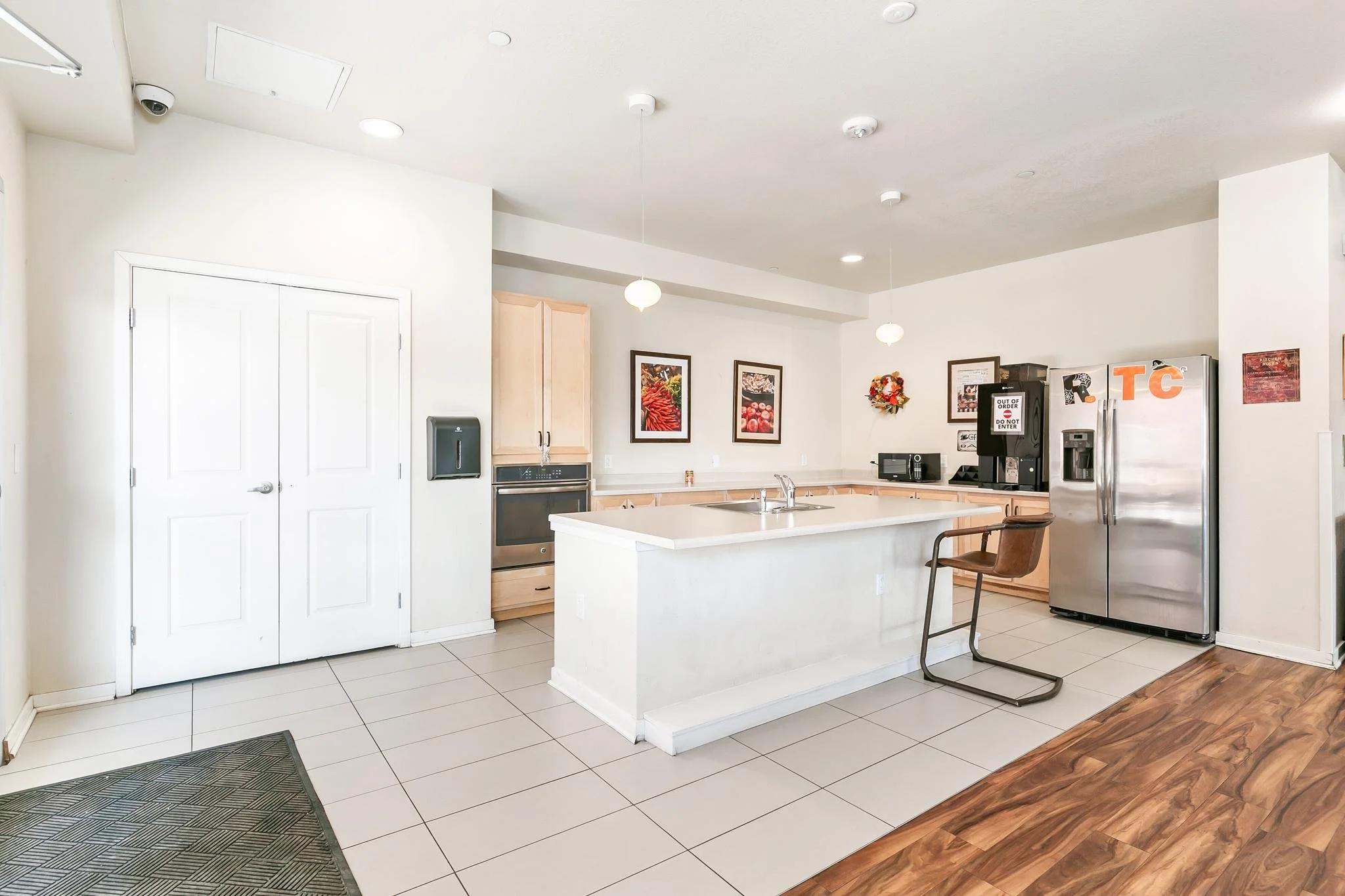 A modern kitchen with white tiled floor, white walls, stainless steel appliances, a kitchen island with a sink, framed pictures on the wall, and a wooden chair.