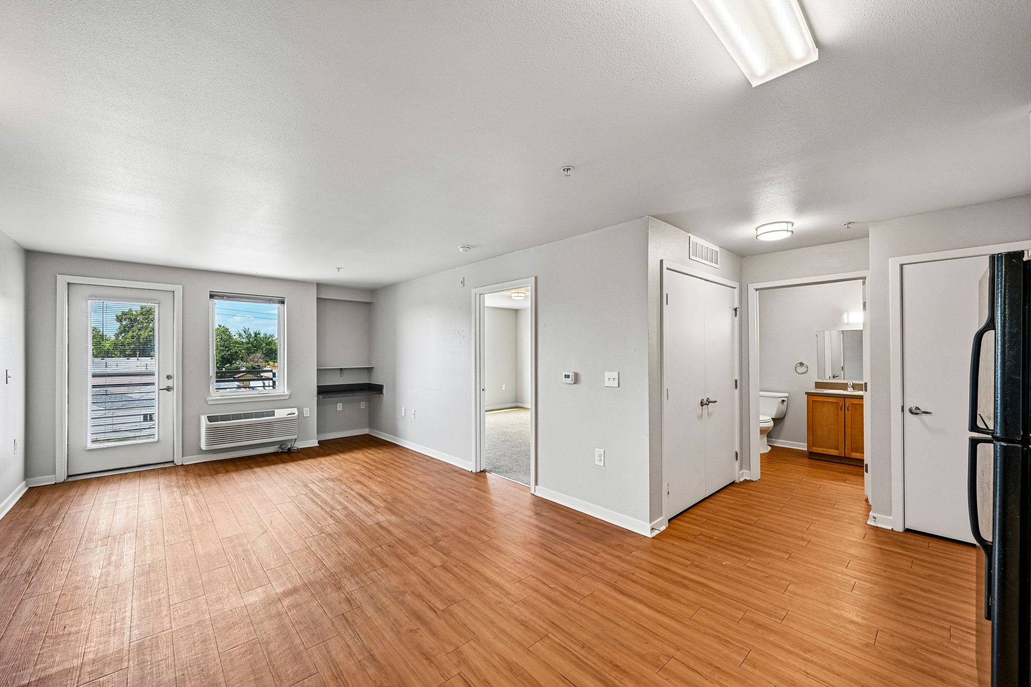 Empty apartment living room with hardwood floors, large windows, white walls, and view into a bathroom and kitchen.