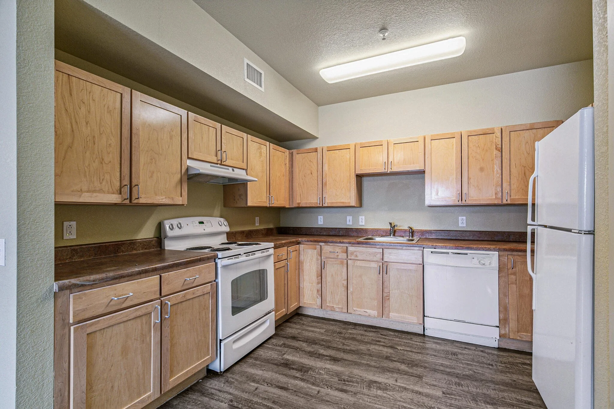 Kitchen with wooden cabinets, white stove, dishwasher, and refrigerator, with beige walls and wood laminate flooring.
