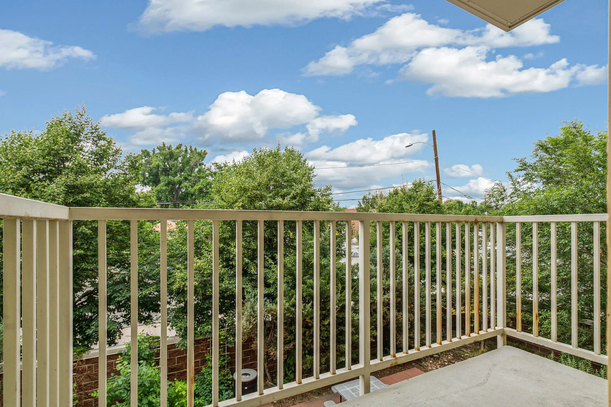 View of a balcony with a beige rail, overlooking green trees and blue sky with scattered white clouds, part of an apartment or house exterior.